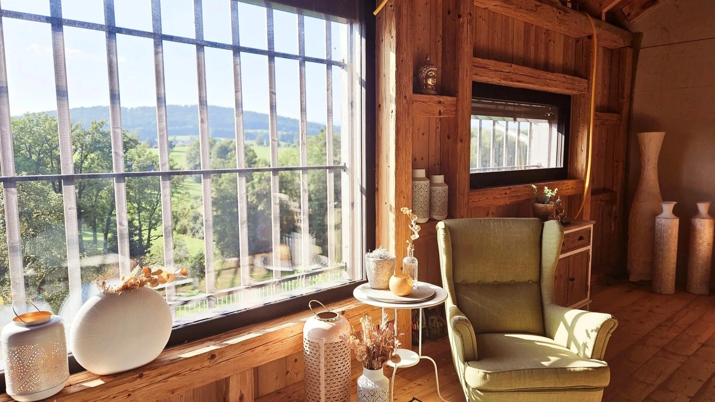 Ein gemütliches Wohnzimmer mit großen Fenstern, Holzwänden und -böden, stilvollen Vasen, Pflanzen und einem grünen Sessel, das Sonnenlicht strömt herein.