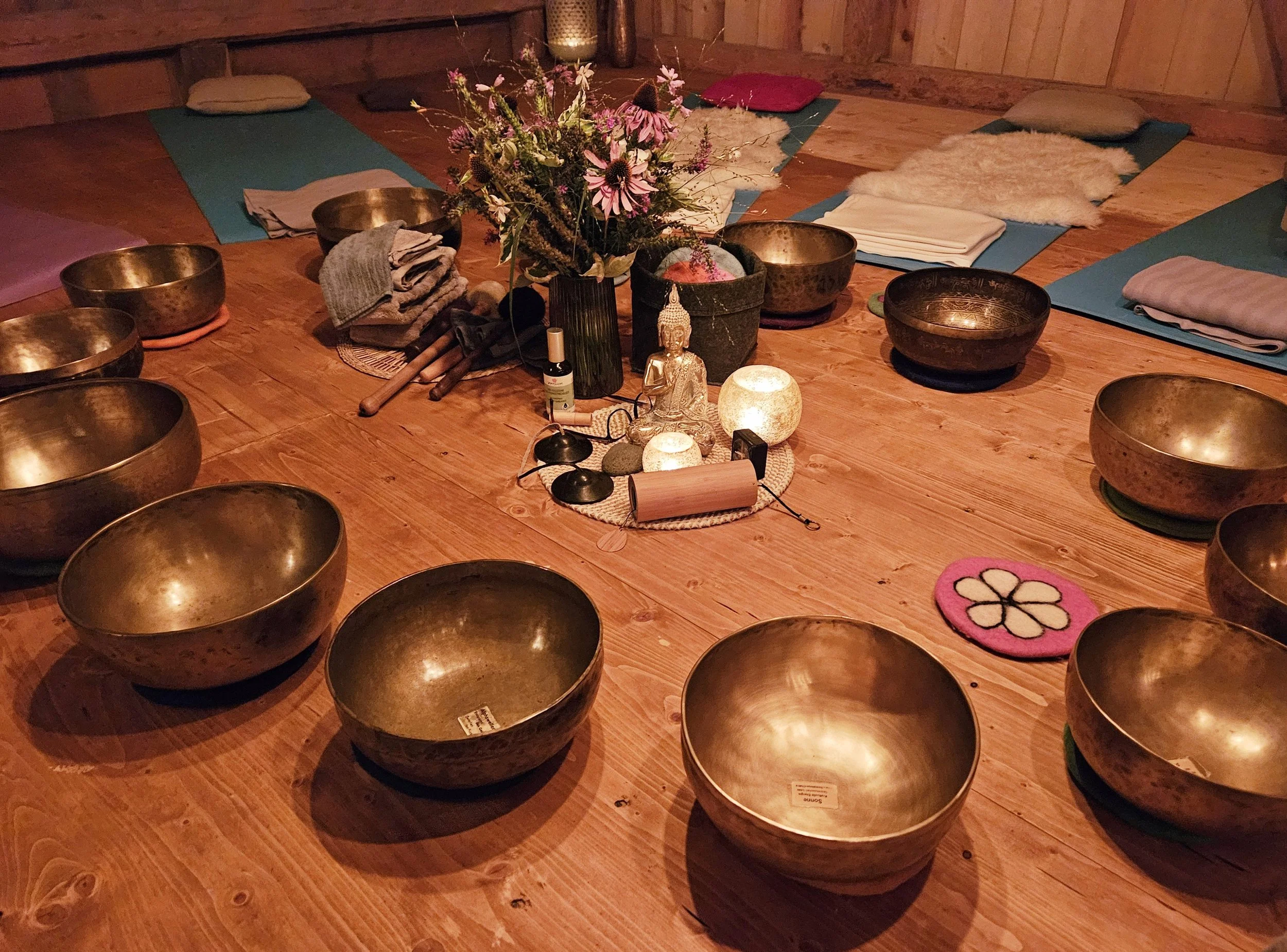 Ruhiger Raum mit tibetischen Klangschalen, Kerzen, einer Buddha-Statue, Blumen und Matten für Meditation auf einem Holzboden.