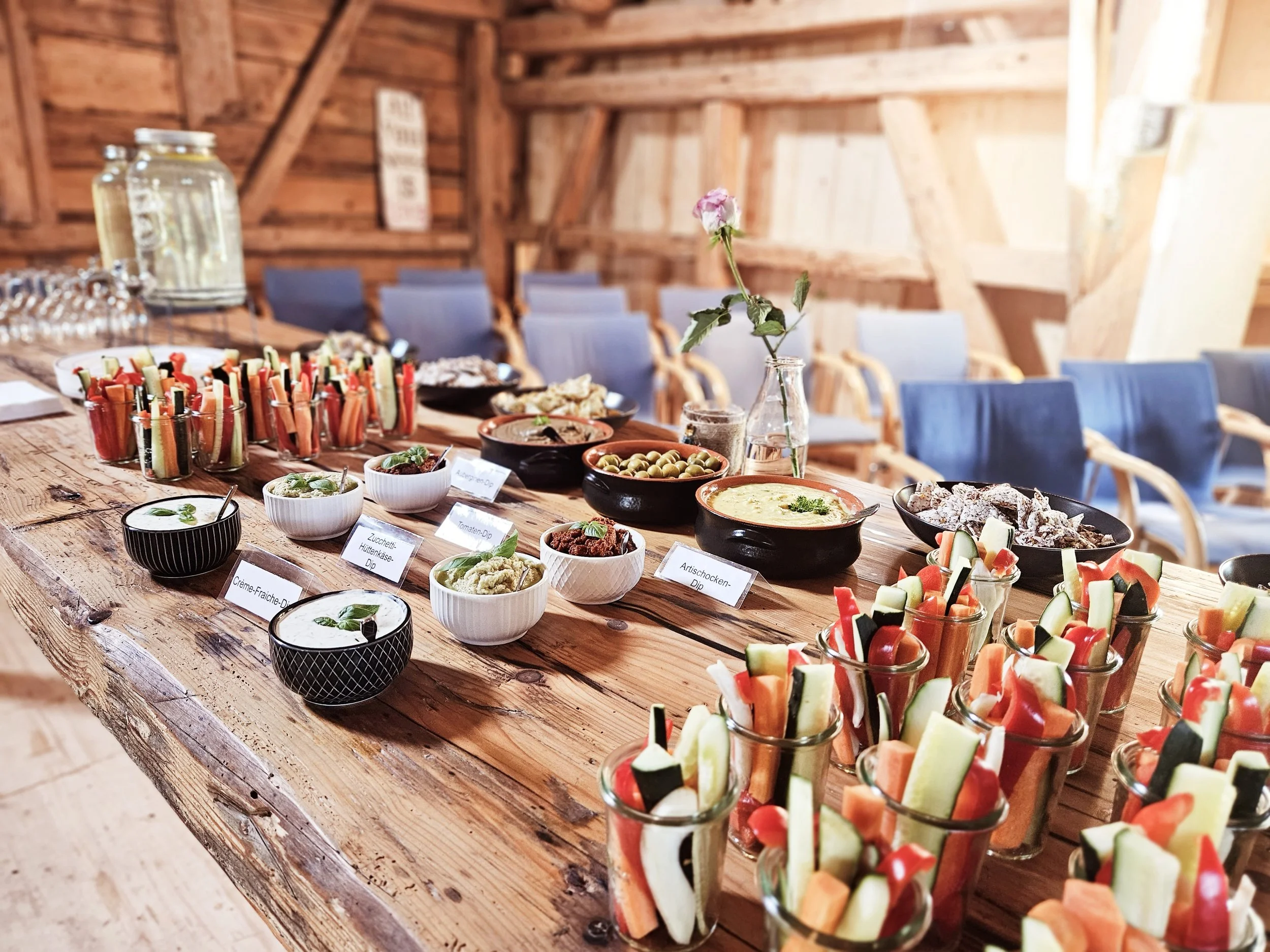 Verschiedene Vorspeisen und Snacks, darunter Gemüsesticks, Aufstriche, Oliven und Dips, auf einem Holztisch in einem rustikalen Raum mit blauen Stühlen.