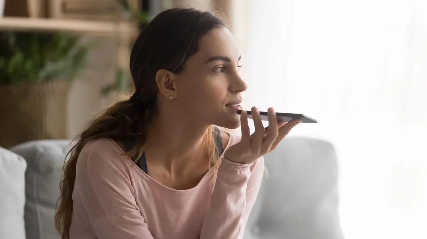 Frau sitzt auf einem Sofa, spricht mit einem Smartphone in der Hand vor dem Ohr, die Umgebung ist hell und wohnlich. Es scheint als ob Sie sich mit jemandem per Sprachnachricht austauscht.