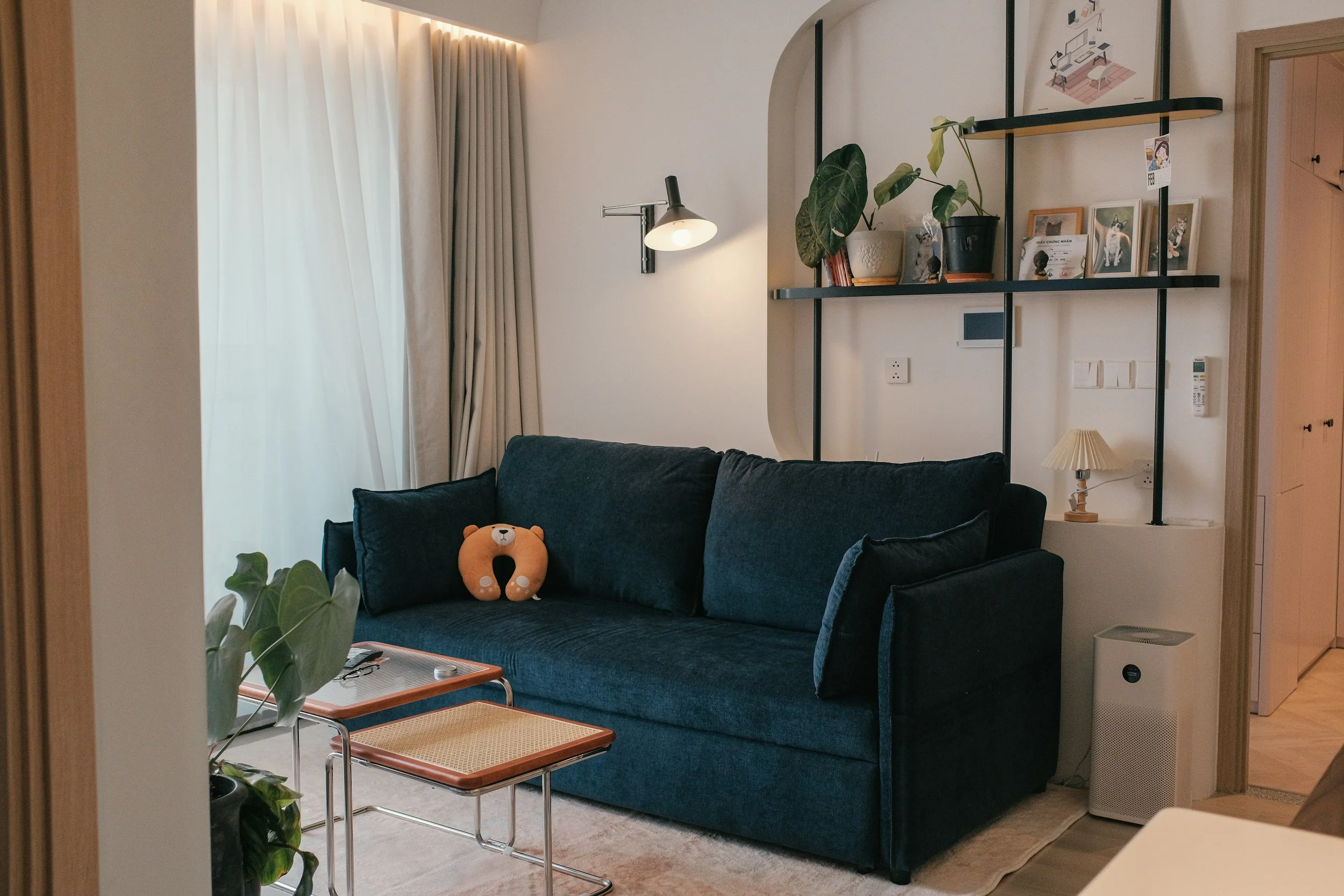 Living room with a dark blue sofa, a plush bear pillow, a small side table, potted plant, and a shelf with pictures and decorations, near a window with beige curtains.