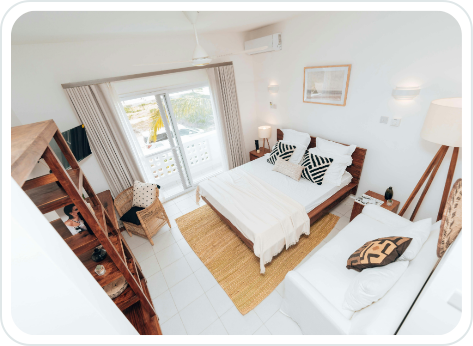 A bedroom with a large bed, a white sofa, and a sliding glass door leading to a balcony. The room has white walls, a wooden shelf, and a wicker chair with cushions. There are framed pictures on the walls and various lamps providing lighting.