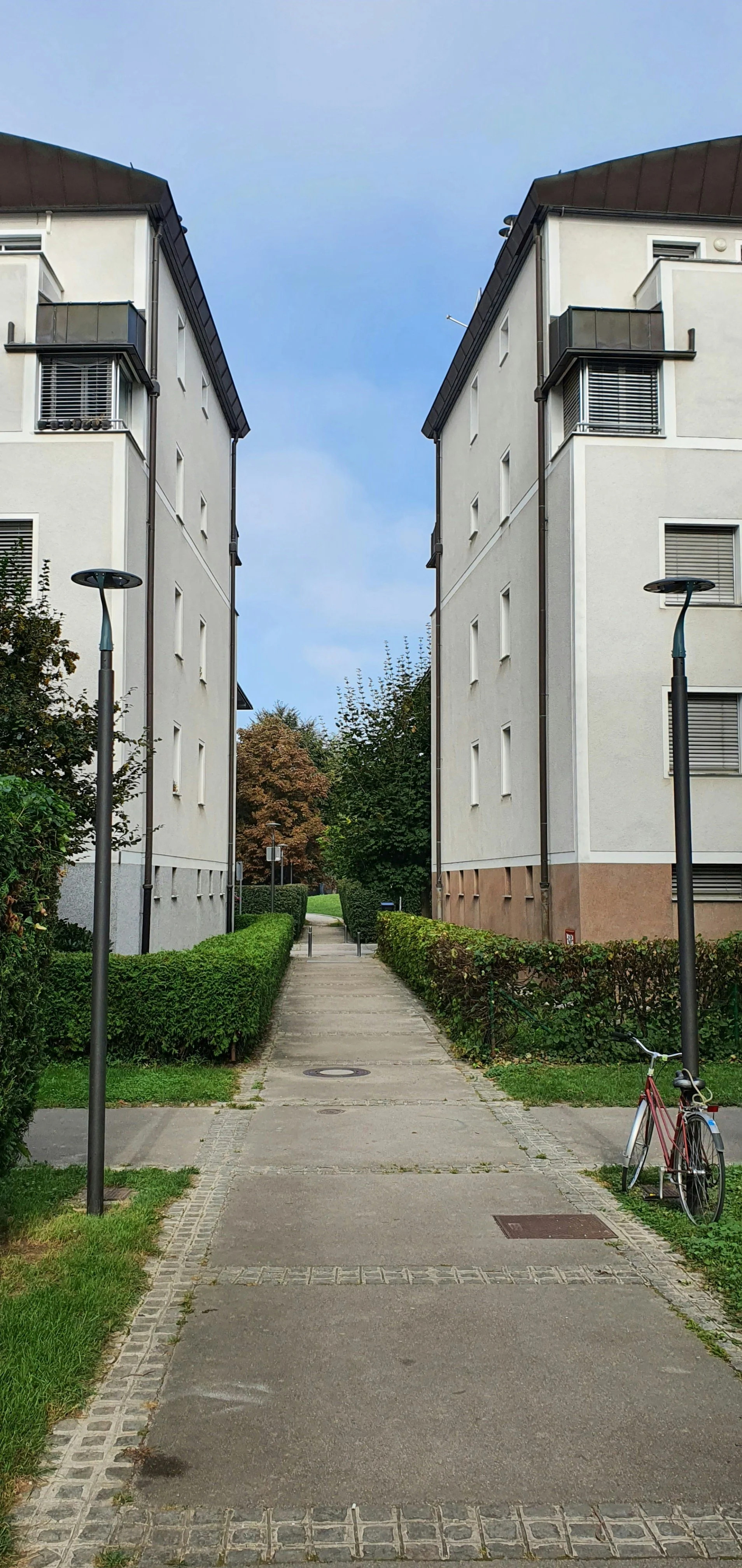 A residential sidewalk between two white apartment buildings with greenery on either side, street lamps, and a bicycle parked near one of the lamps.
