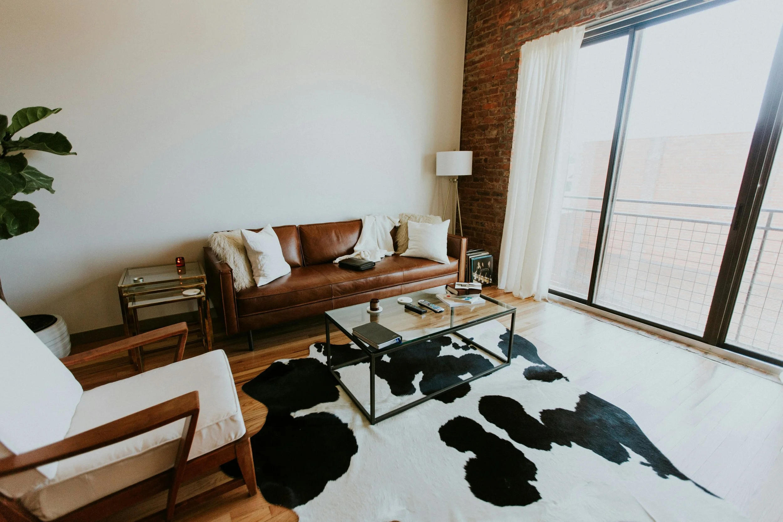 Living room with a leather couch, white armchair, glass coffee table, cowhide rug, large window with curtains, exposed brick wall, and a green plant.