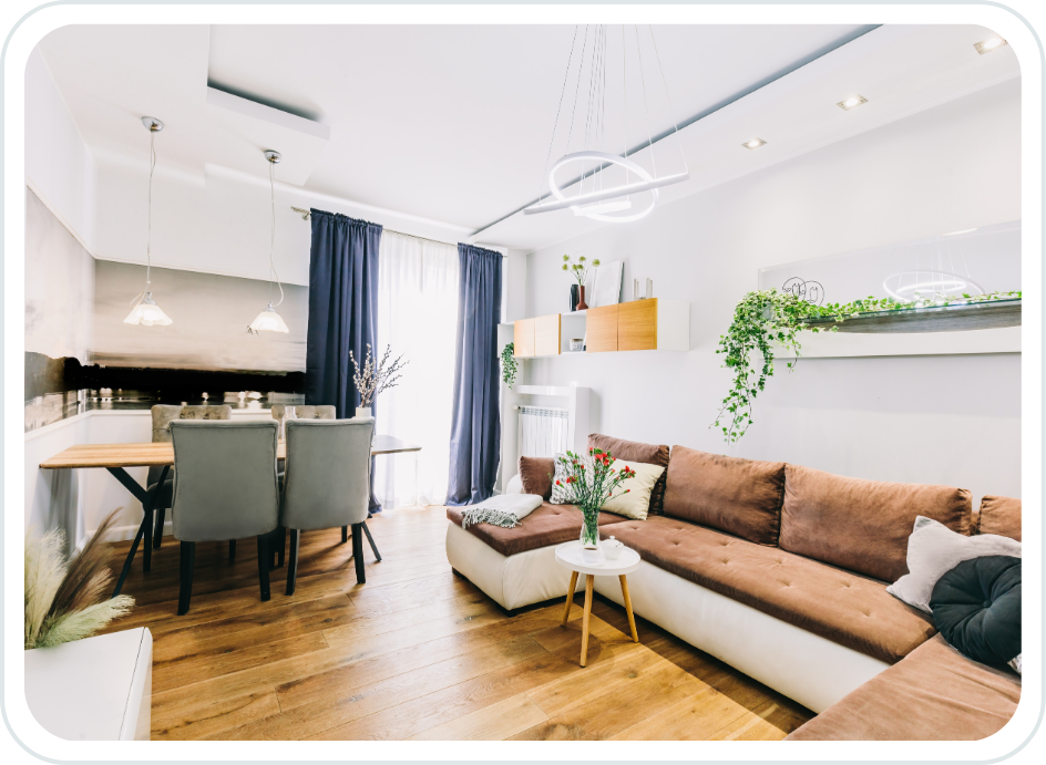 Modern living room with brown and white sectional sofa, small white coffee table with a vase of flowers, wall-mounted shelves with plants, dining area with four gray chairs and a wooden table, large window with dark curtains, and contemporary lighting fixtures.