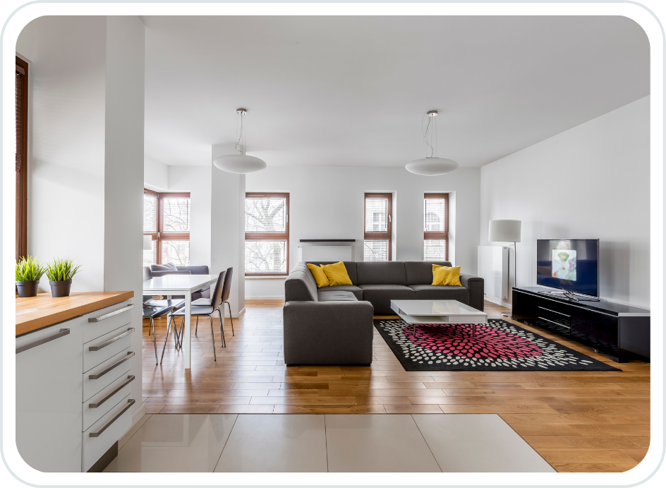 Modern open-concept living room with a gray sectional sofa, yellow pillows, white coffee table, black TV stand with a flat-screen TV, and a patterned rug. Adjacent dining area with a table and chairs, white walls, hardwood flooring, and multiple windows.