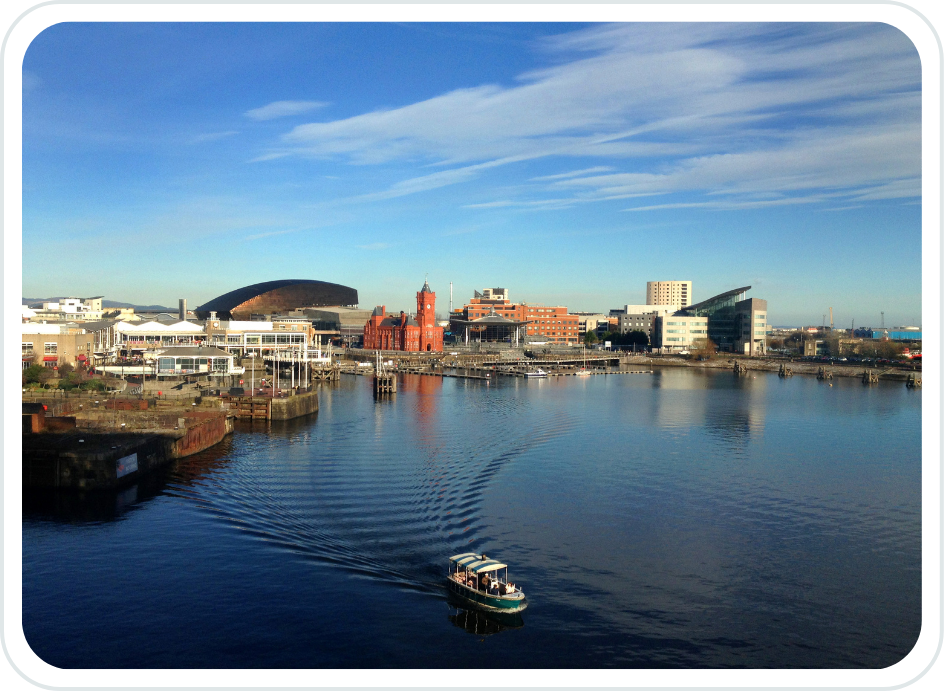 A city skyline reflecting on a calm body of water, with a small motorboat moving across the water. The city includes modern and historic buildings, with a clear blue sky overhead.