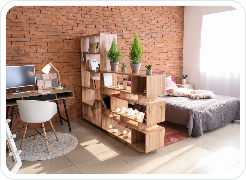 A cozy bedroom with a brick wall, a bed with gray bedding and pink pillows, a wooden bookshelf dividing the space, a desk with a computer and lamp, and a window with white curtains.