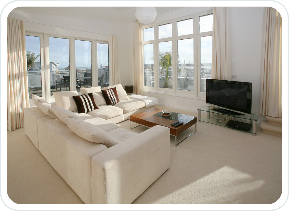 Bright living room with white sectional sofa, black and white throw pillows, large windows with curtains, and a flat-screen TV on a glass stand.