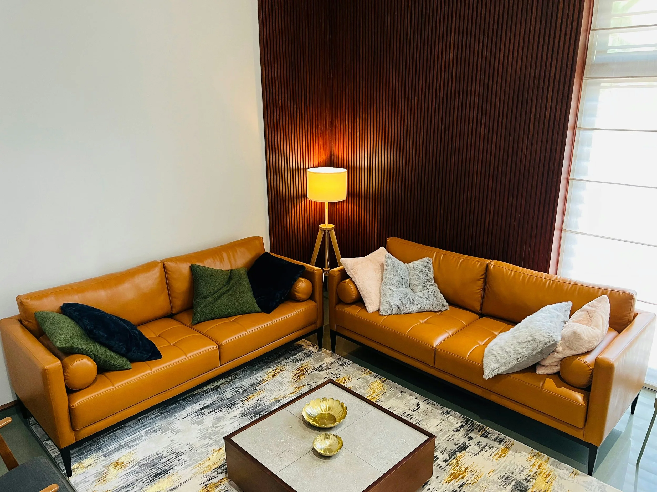 Living room with two brown leather sofas, a wooden coffee table with gold bowls, a tall floor lamp with a white shade, and decorative pillows in various colors and textures. One wall is white, and the other is wood-paneled. There are large windows with sheer curtains.