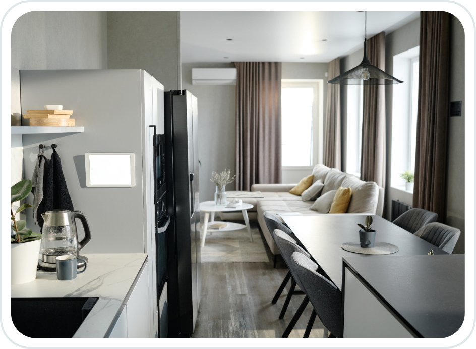 Modern living room and kitchen with gray walls, large windows with brown curtains, gray sofa with yellow and gray pillows, dining table with gray chairs, and a refrigerator with a water pitcher and mug on the kitchen counter.