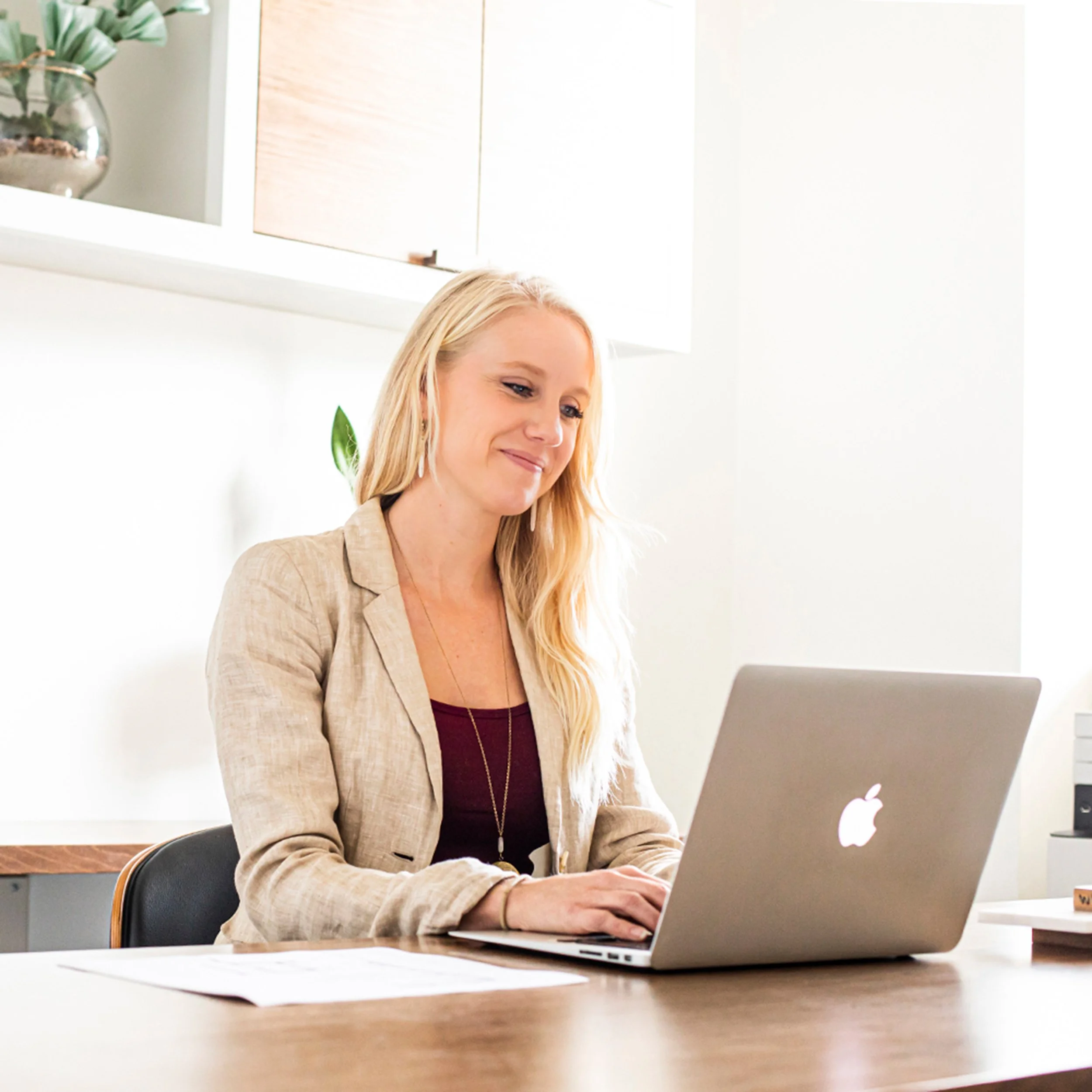 A woman with long blonde hair working on a silver MacBook in an office setting