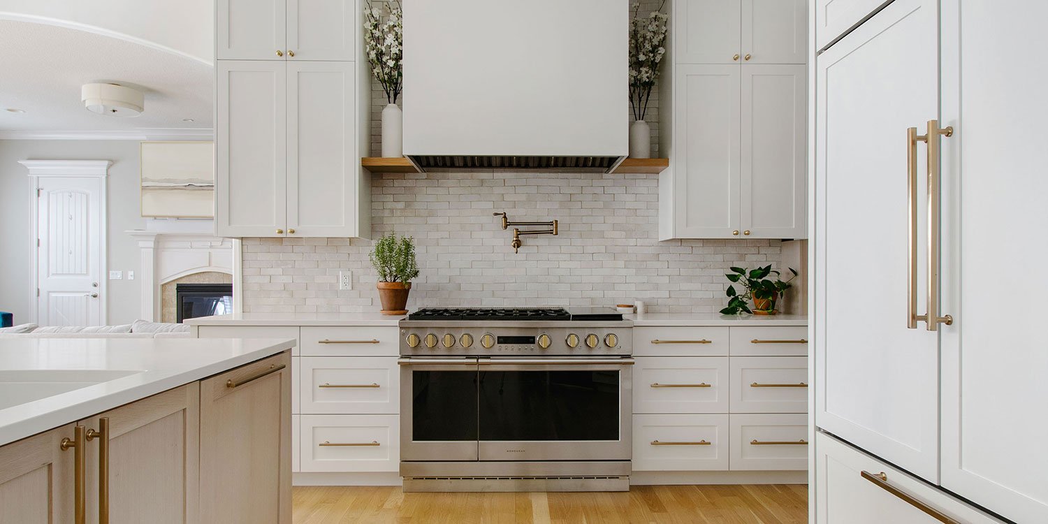 Completed luxury kitchen renovation in Kansas City by Erica Kay Design & Remodeling, featuring a professional 48-inch range, a custom white hood with wood mantle detail, a light grey zellige tile backsplash, and elegant brass hardware.