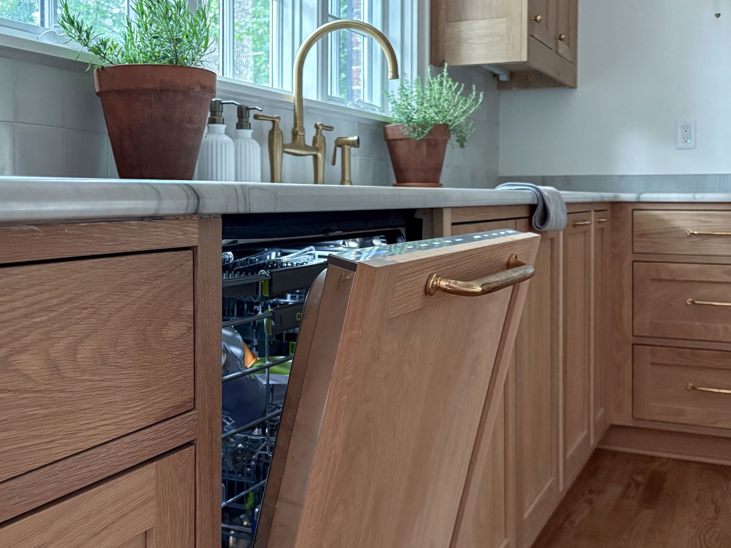 Cabinet-front dishwasher beneath a honed stone countertop, beside a kitchen sink with a brass faucet and white oak cabinetry.