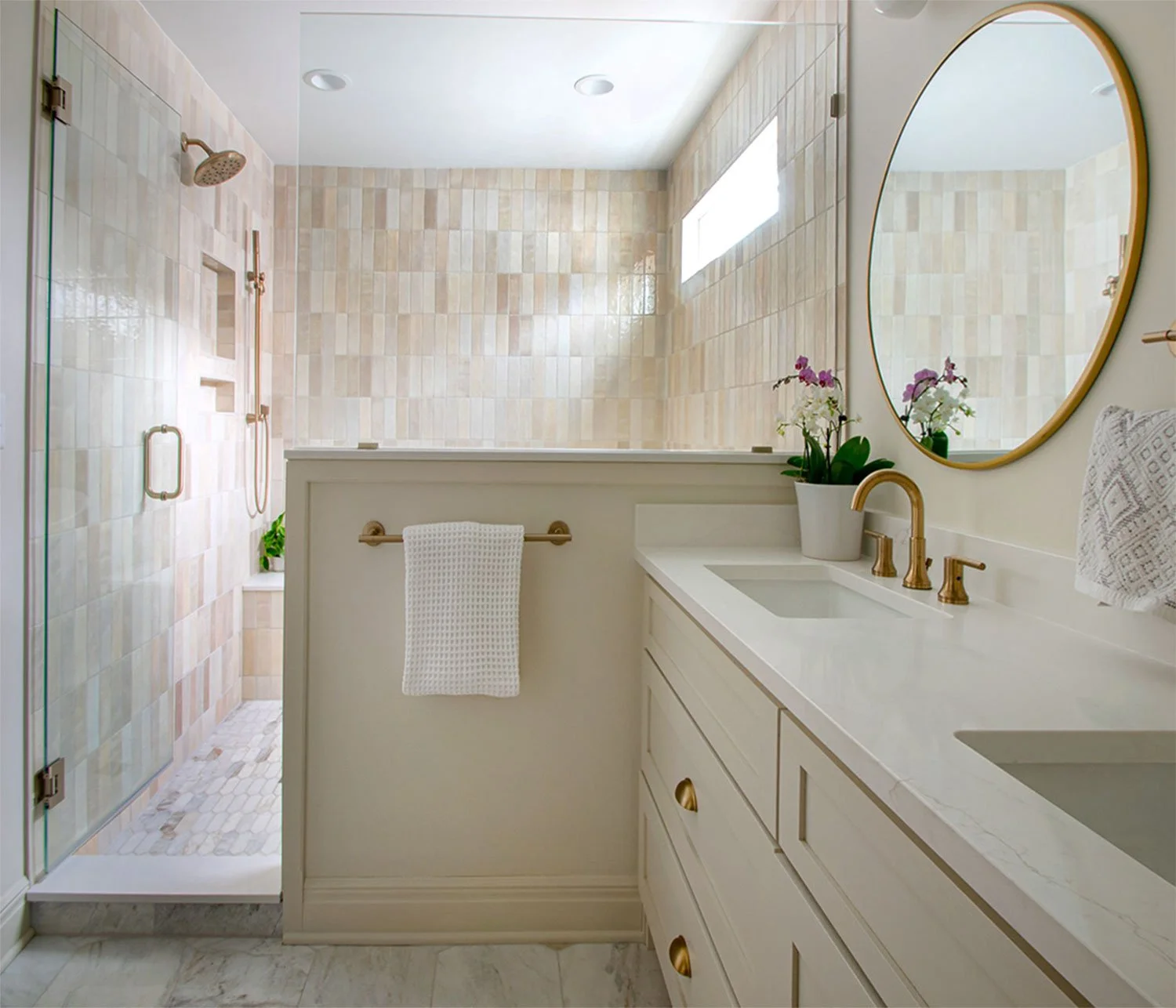 Luxury primary bathroom remodel in Kansas City by Erica Kay Design & Remodeling featuring a warm beige double vanity with gold hardware, round brass mirrors, and a walk-in shower with vertical handmade tile and marble mosaic flooring.