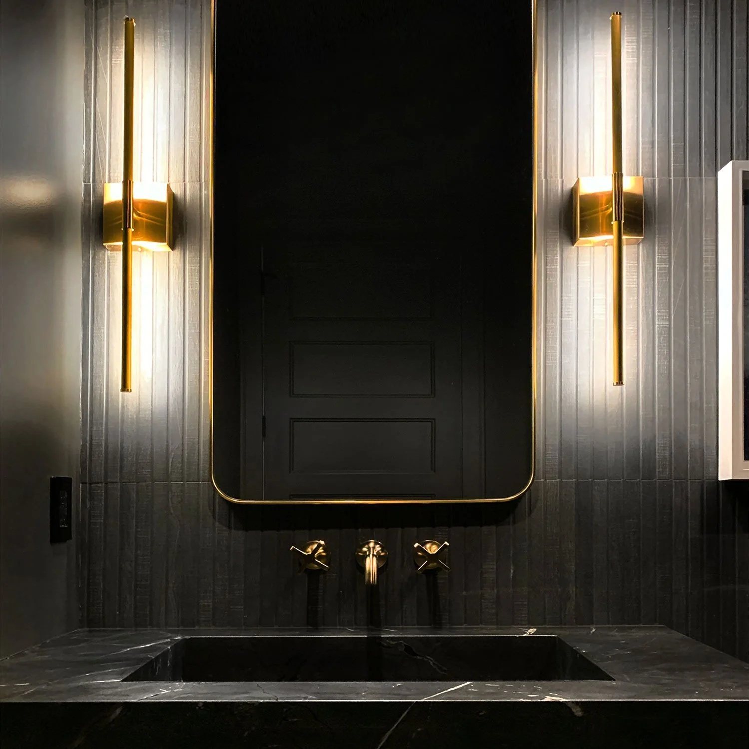 Modern luxury bathroom design in Kansas City by Erica Kay Design & Remodeling featuring an integrated black quartzite mitered sink and vanity, dark vertical fluted wall tile, a gold-rimmed mirror, and elegant brass wall sconces.