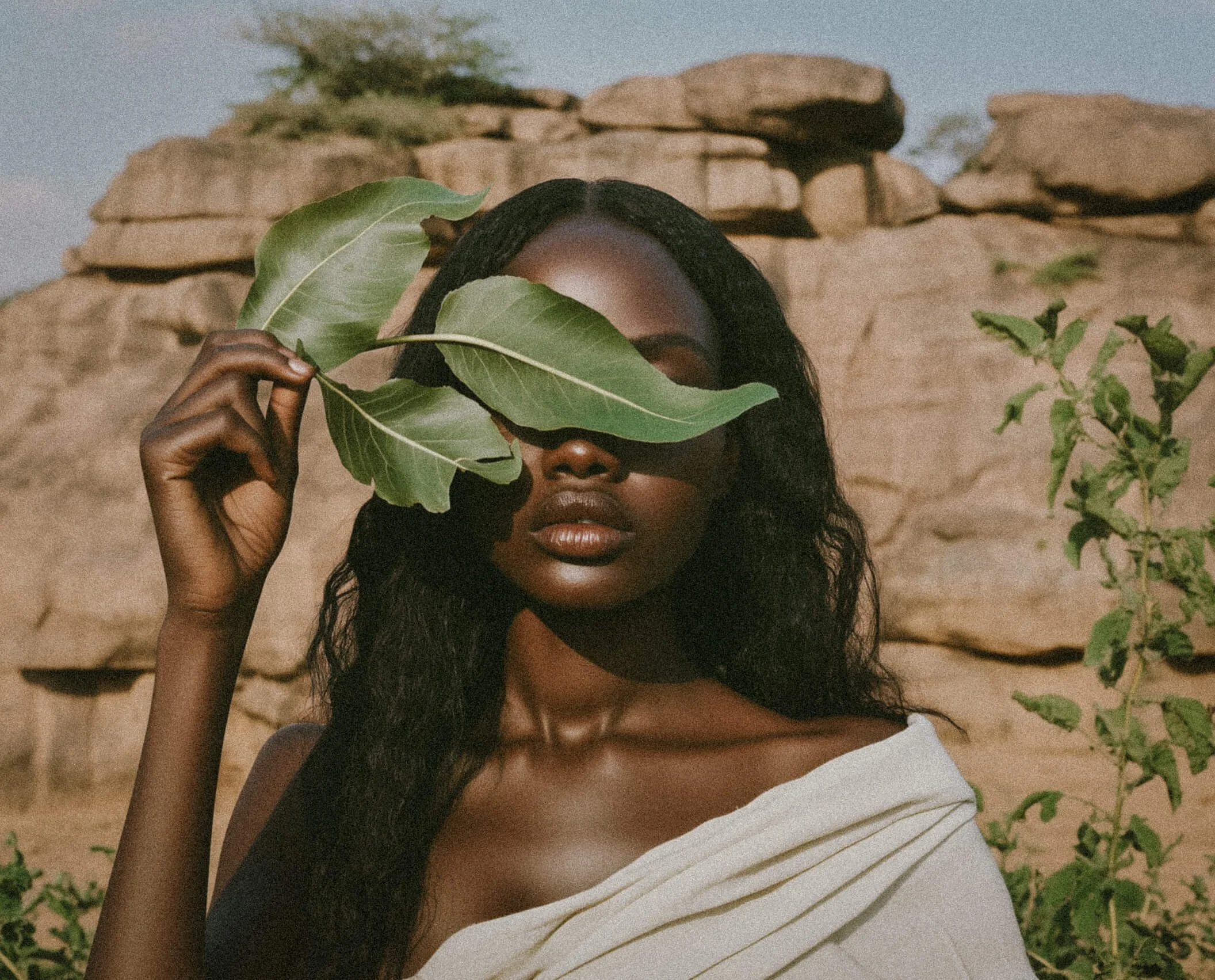 A woman with dark skin and long black hair holds a large green leaf in front of her face, obscuring her eyes, against a rocky outdoor landscape.