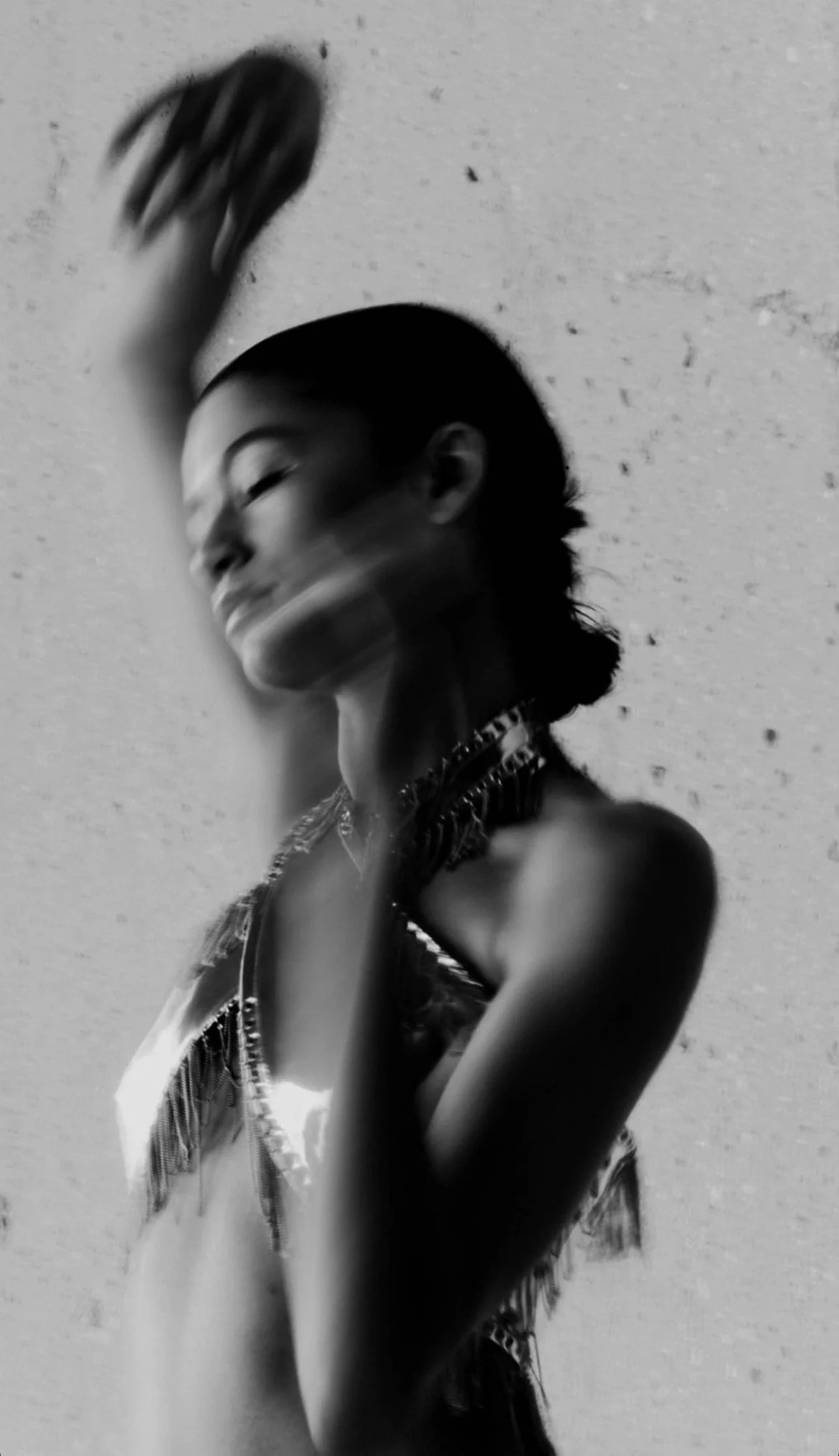 A young woman with short hair and layered necklaces poses against a textured wall, smiling and raising her arm, in black and white.