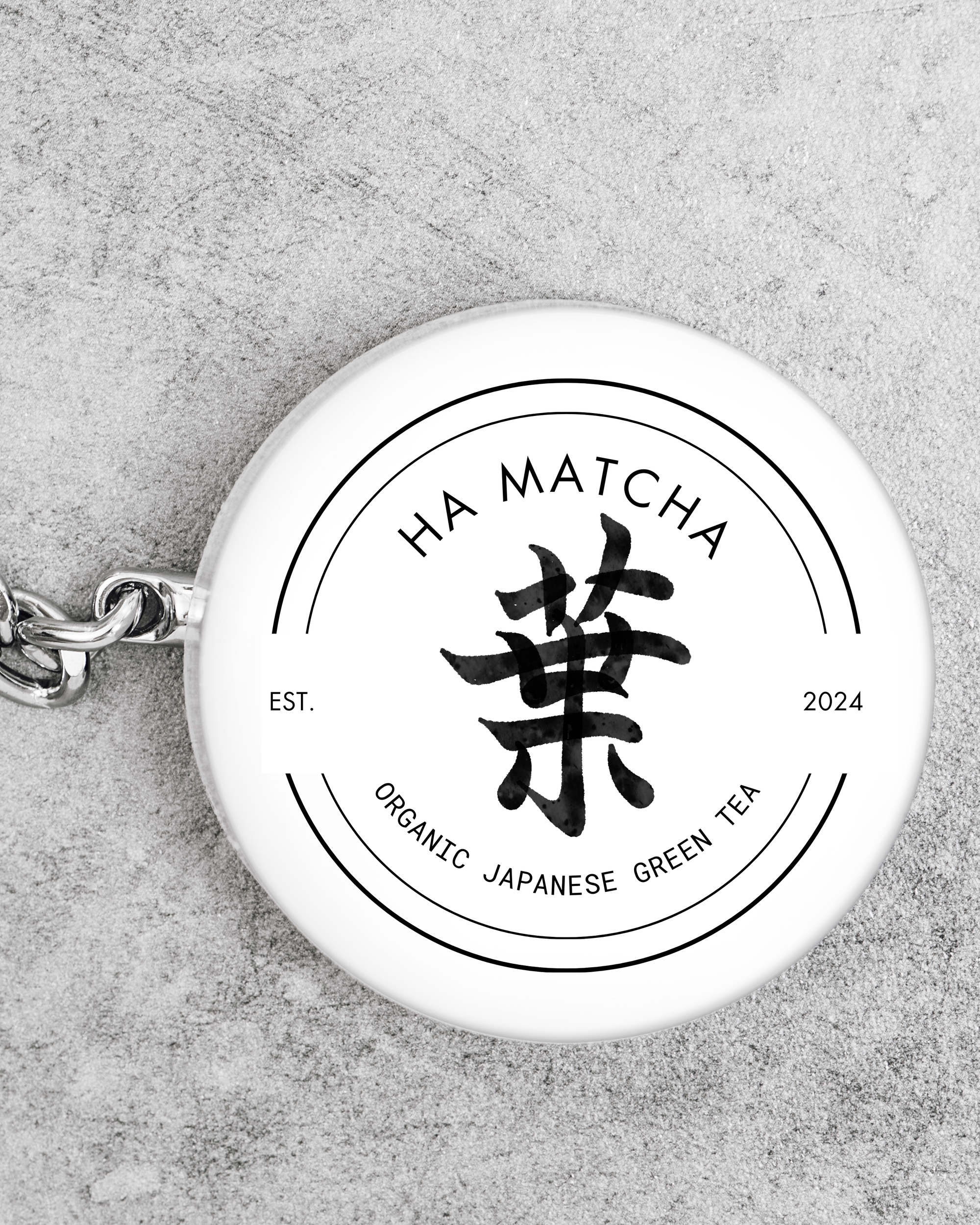 Llavero blanco con diseño de té de hoja verde orgánico japonés, marca 'Ha Matcha', Est. 2024, sobre superficie de piedra gris.