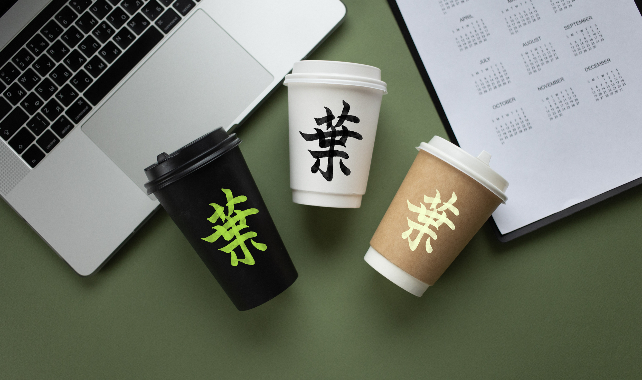 Tazas de café con tapa, una negra, una blanca y una marrón, con caracteres chinos en ellas, sobre una mesa verde junto a una laptop y una agenda con calendario