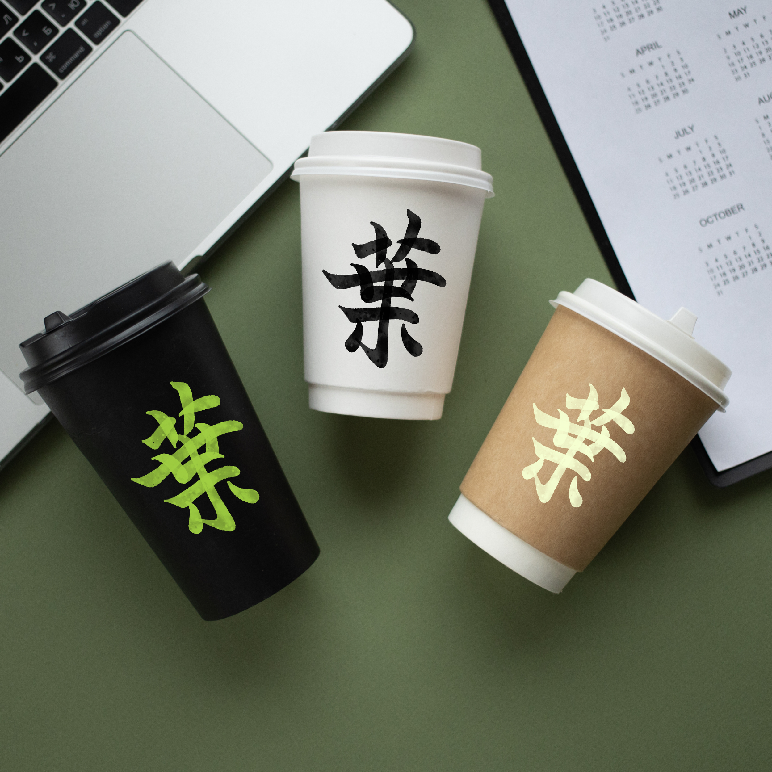 Tres vasos de café con tapa de plástico, uno negro, uno blanco y uno color kraft, con la misma letra en chino impresa en ellos, sobre una mesa con una laptop y un cuaderno a la derecha.