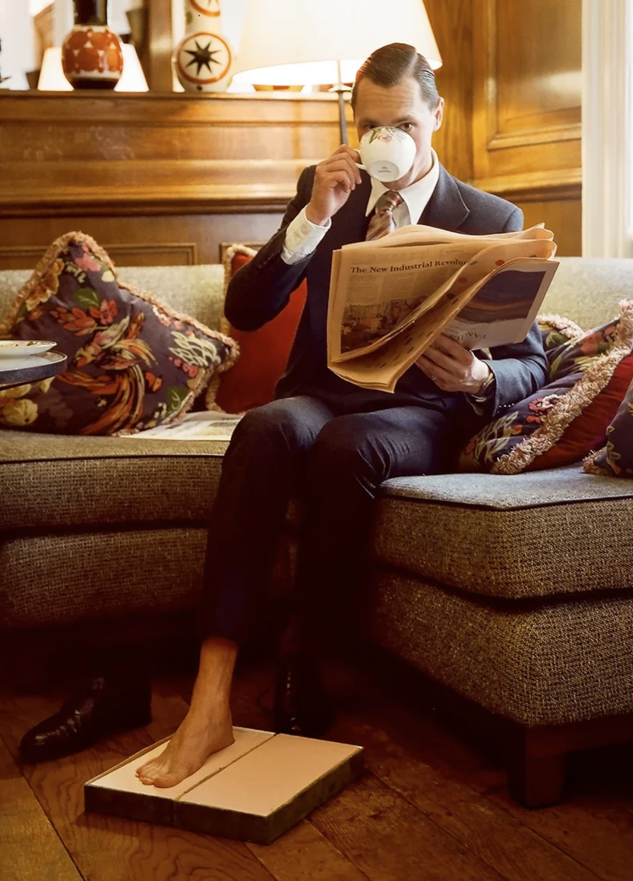 A man sitting on a sofa, drinking from a teacup and reading a newspaper, with one foot on a wooden footstool and the other on a foot measuring device, surrounded by decorative pillows and wooden paneling.