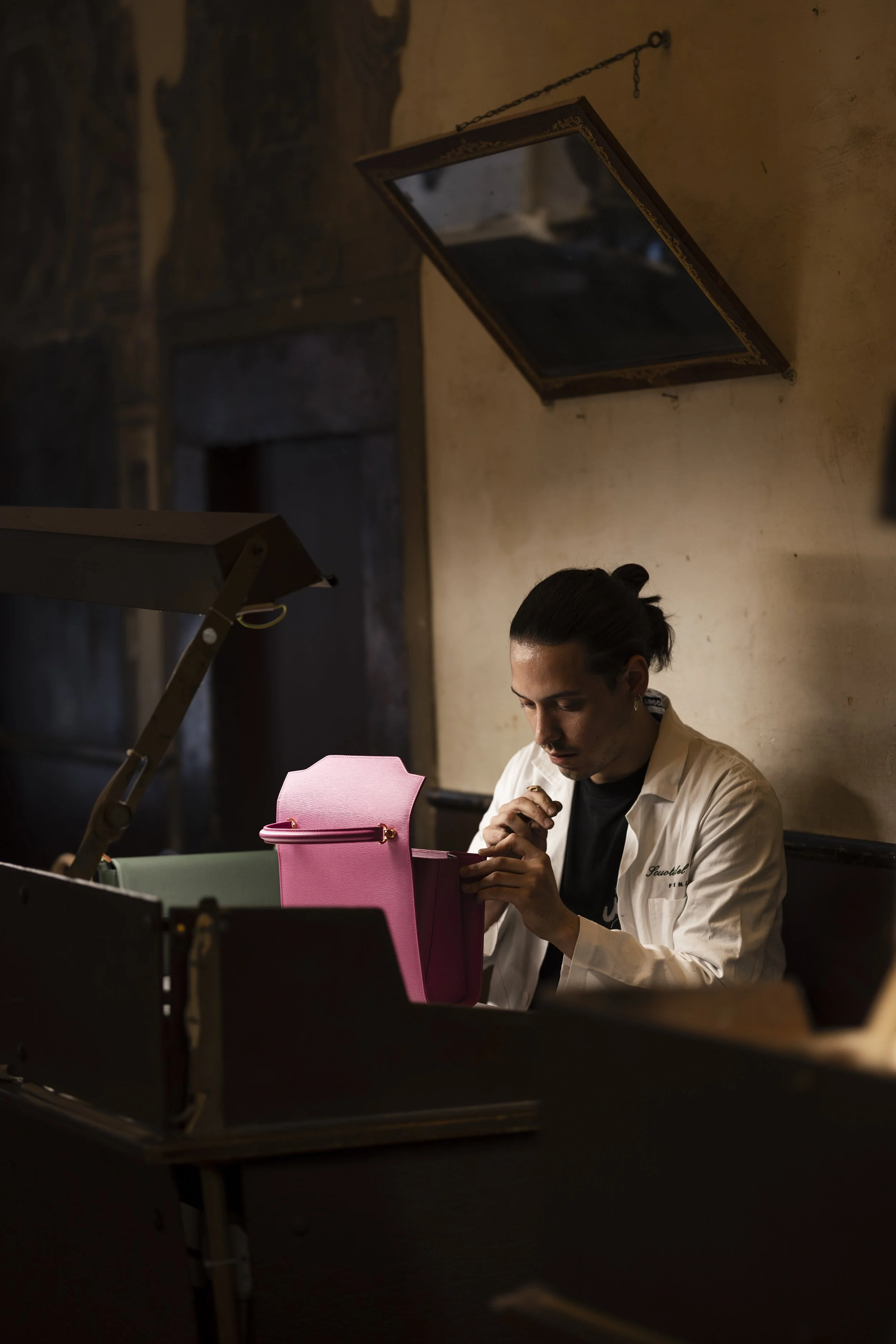 A man with dark hair tied back, wearing a white chef's coat, is sitting at a table in a dimly lit room, looking intently at a pink purse in front of him.