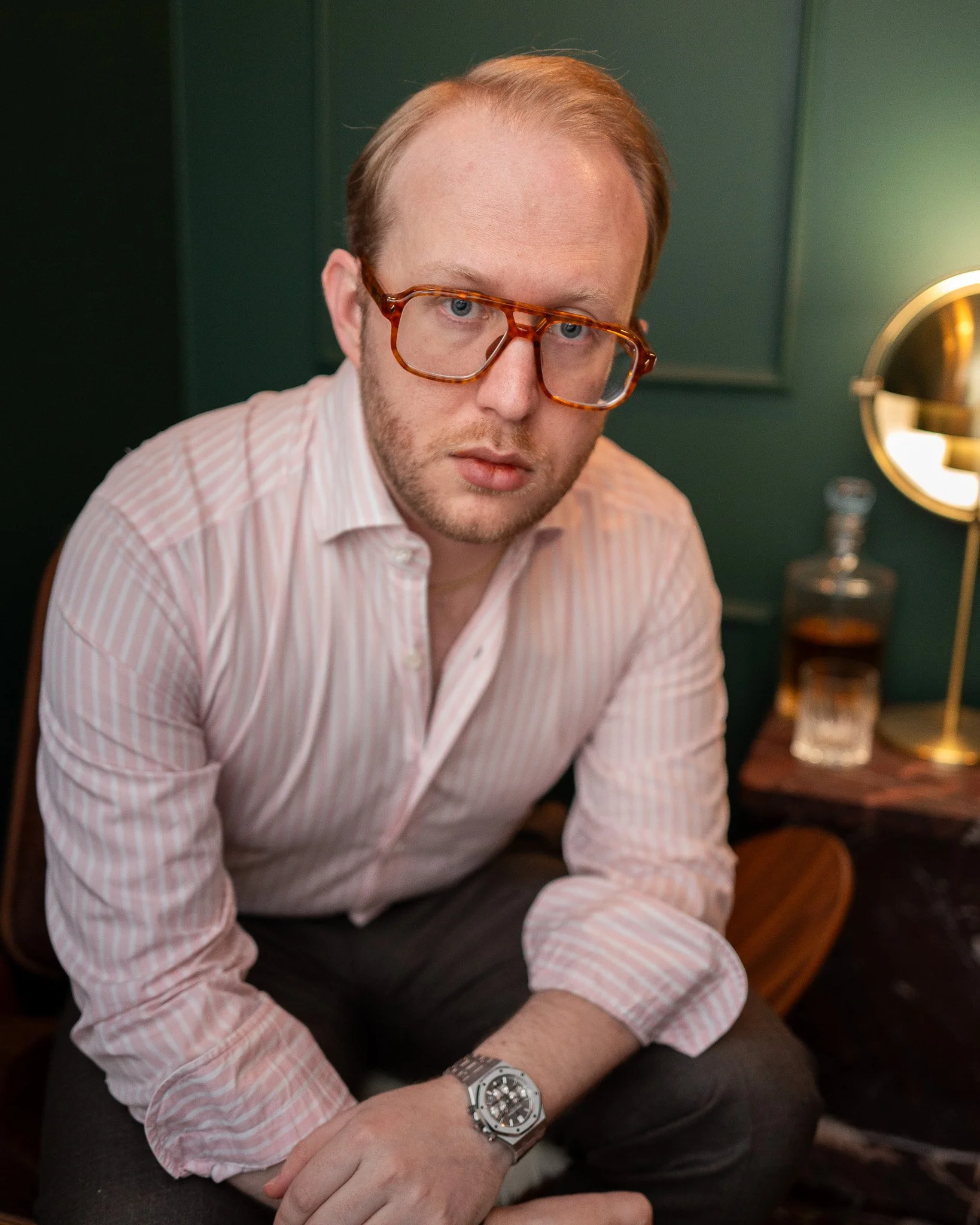 A man with fair skin, red hair, and blue eyes is sitting indoors, wearing a pink and white striped shirt, glasses, and a large silver watch on his left wrist. He appears to be looking directly at the camera. The background includes a green wall and a round mirror with a brass frame, along with some bottles and picture frames on a wooden surface.