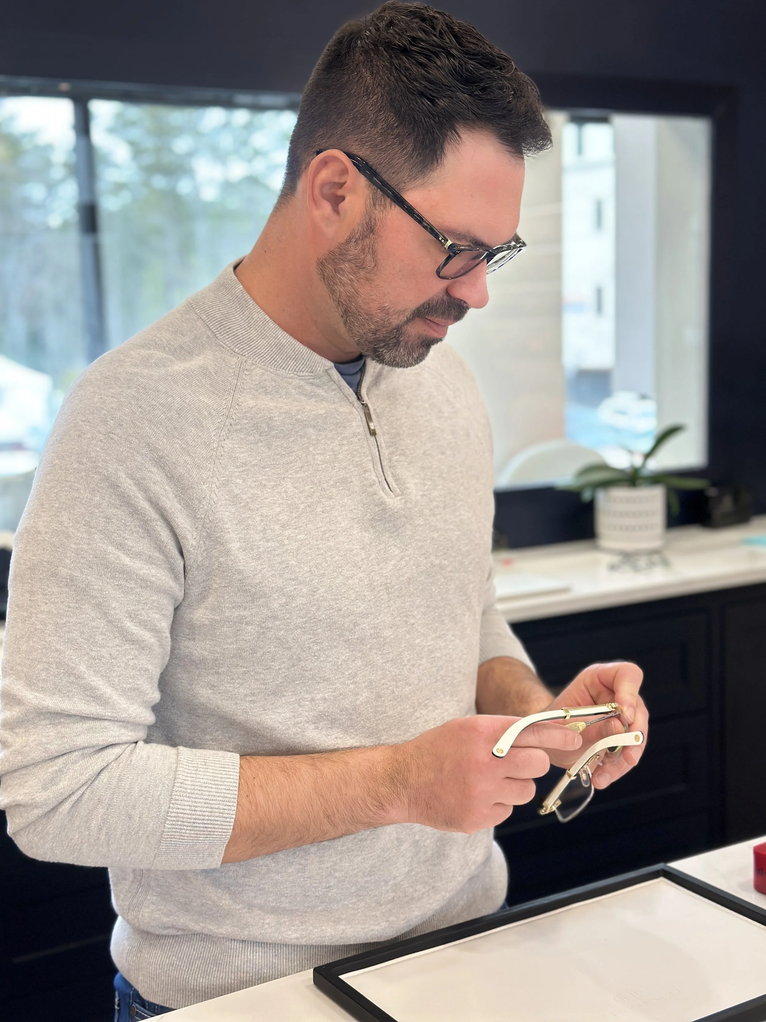 A man with short dark hair, beard, and glasses is examining a pair of white and gold sunglasses, standing in a room with large windows and a plant in the background.