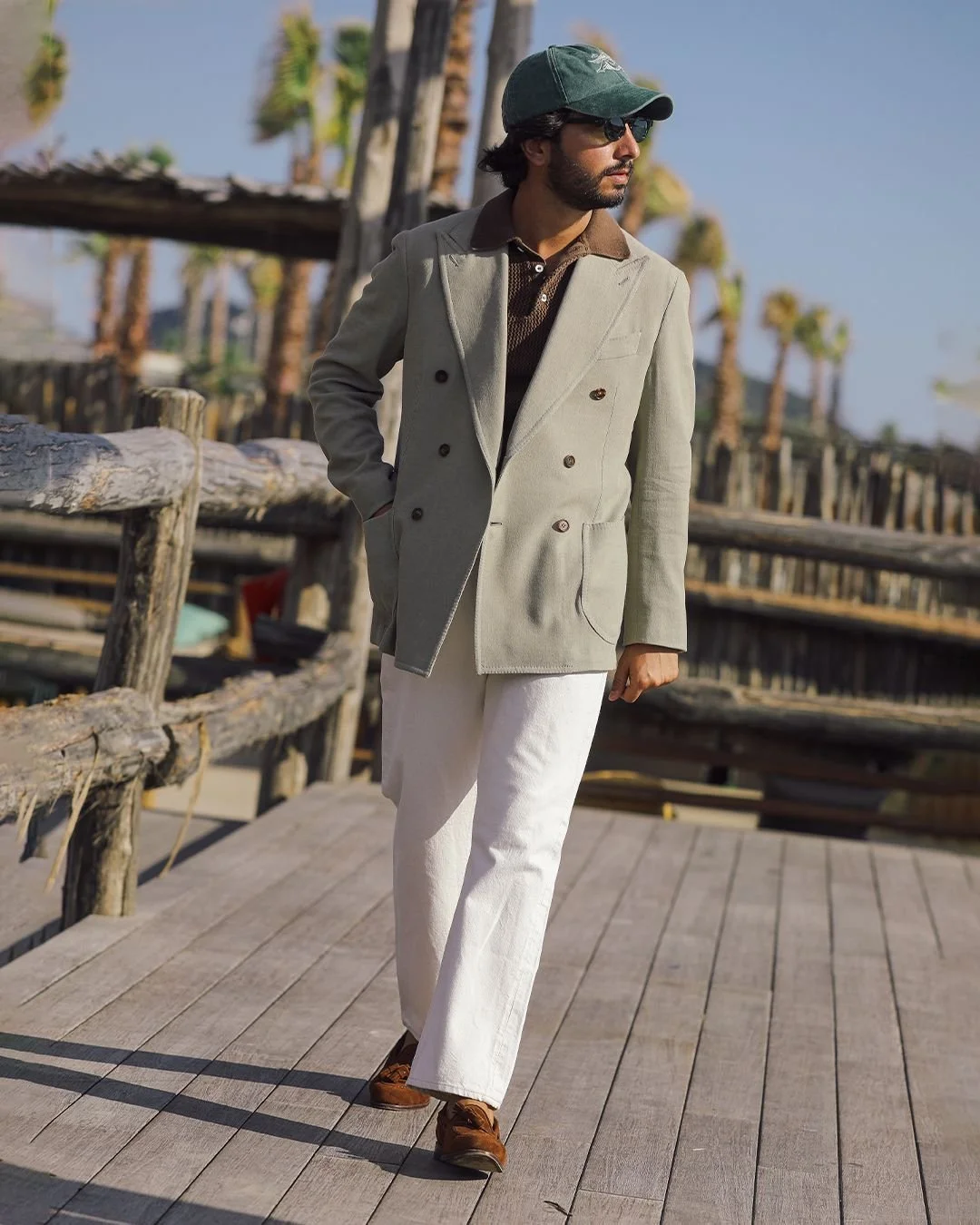 Man in beige double-breasted blazer, brown shirt, white pants, brown shoes, green cap, and sunglasses walking on a wooden pier with palm trees in the background.
