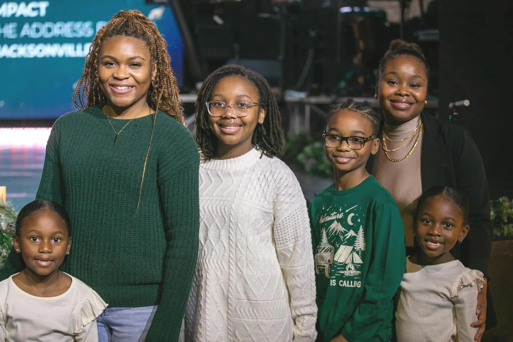 A group of six women and children posing together indoors, smiling. They are dressed casually, with a dark background and a blue screen or banner in the back.