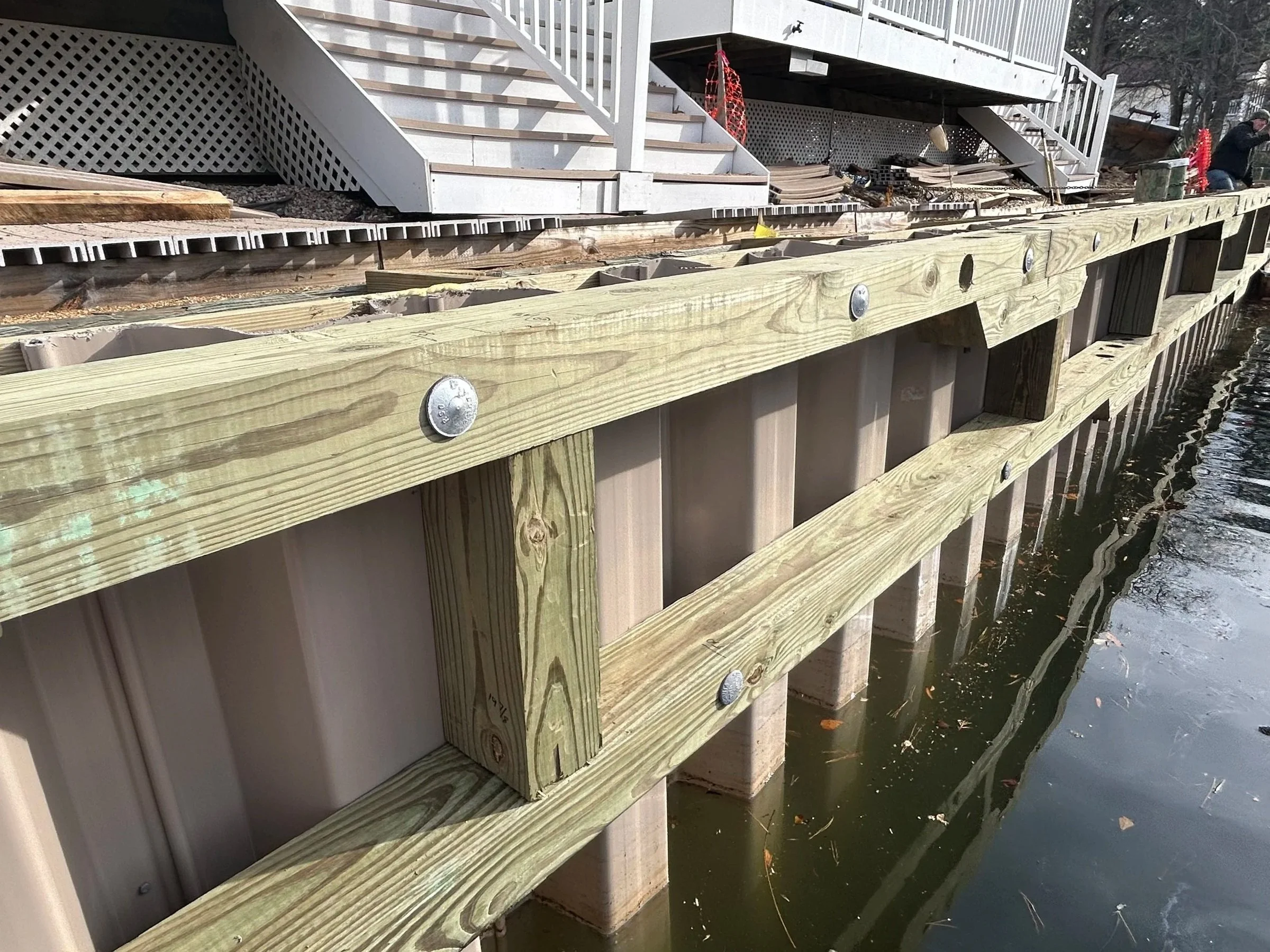 Close-up of a newly constructed wooden dock with screws, extending over water with a house in the background.