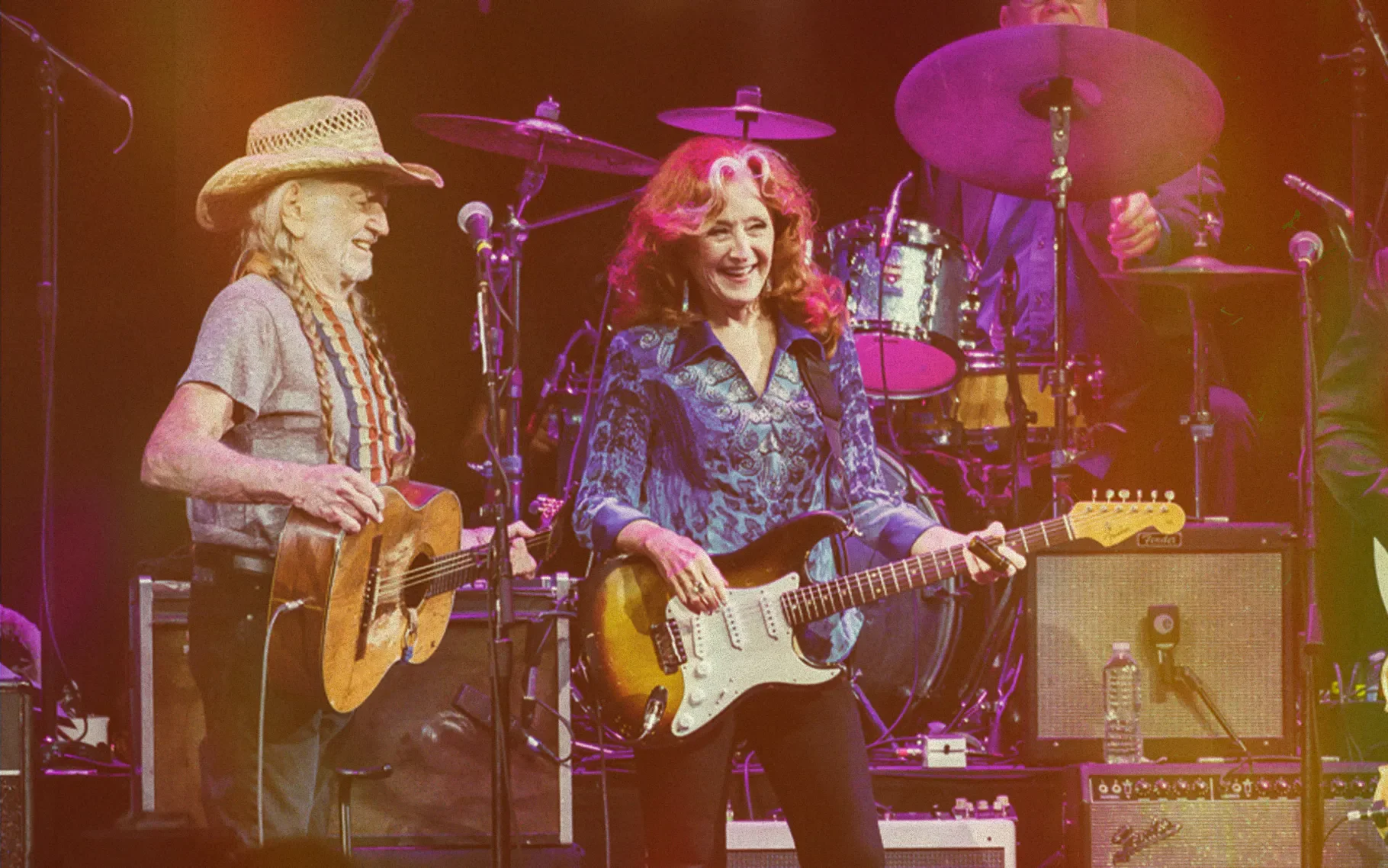 Two women playing electric guitars on stage, smiling, with a band and drum set in the background.