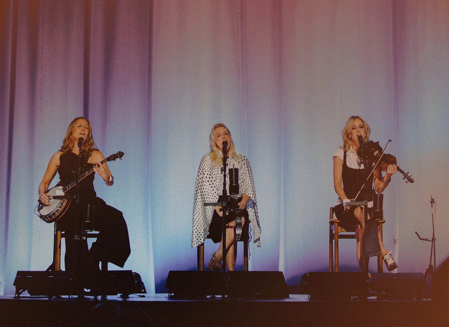 Three women performing on stage, singing into microphones; the woman on the left is playing a banjo, the woman on the right is playing a violin, and the woman in the center is singing with a polka dot shawl.