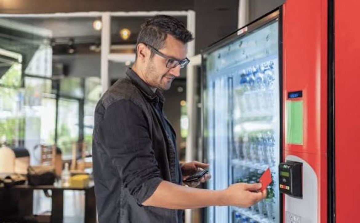 A man with glasses purchasing a snack from a vending machine in a modern cafe.