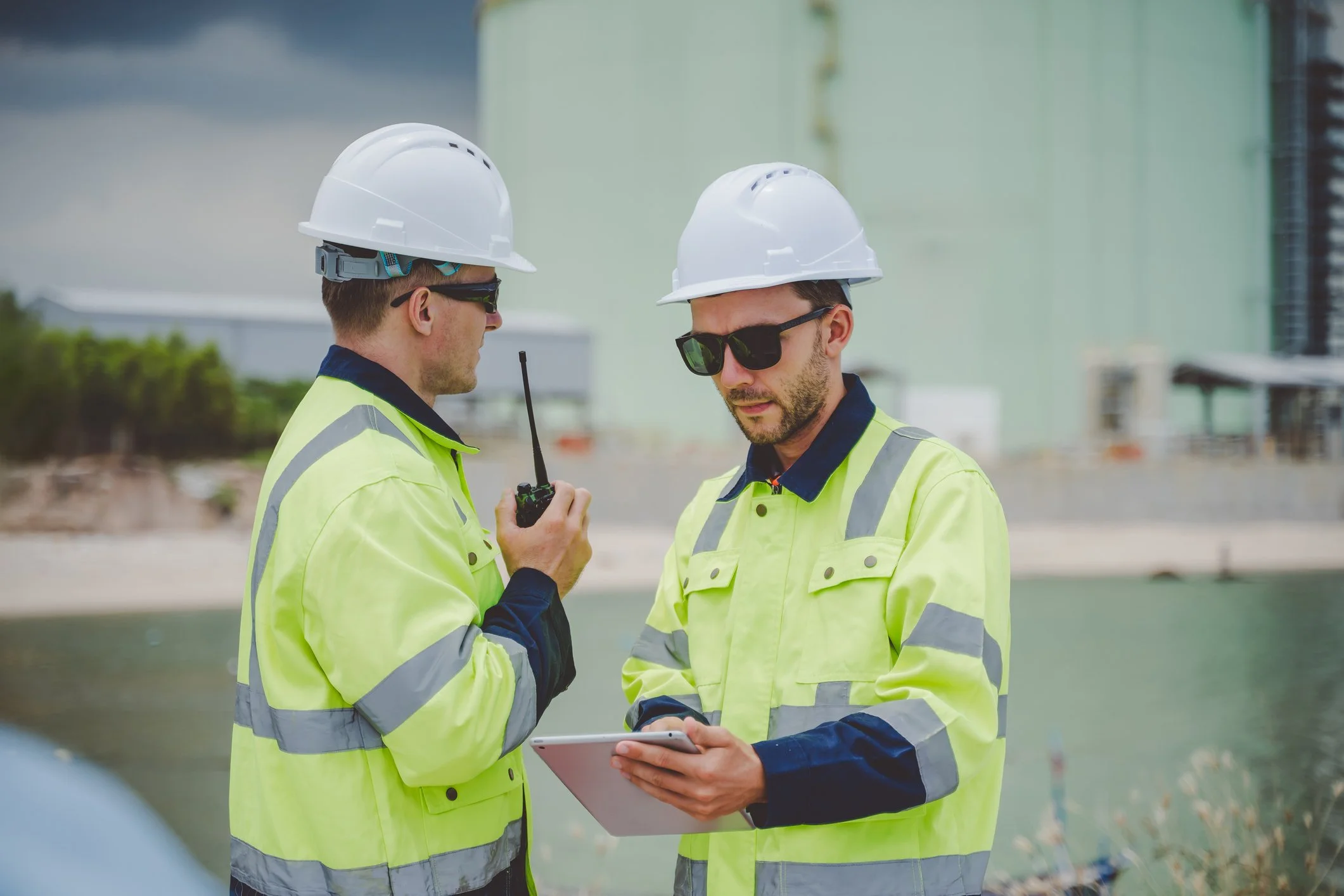 Two construction workers wearing white hard hats and yellow safety vests standing outdoors; one holding a walkie-talkie, the other holding a tablet, having a discussion.