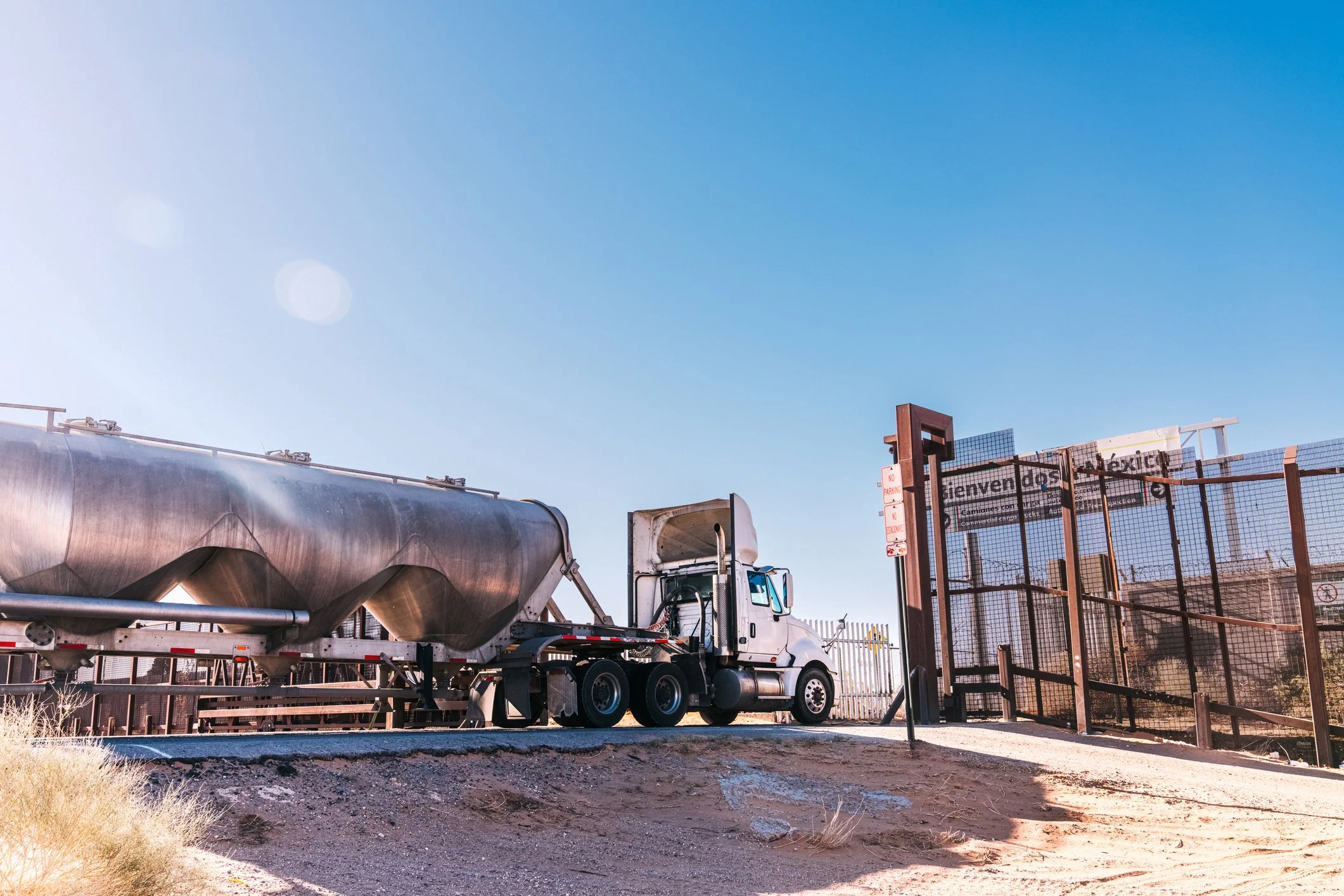 A white semi-truck with a large metal cylindrical tanker trailer parked near a fenced perimeter with metal gates and a sign that says "No Parking." The sky is clear and blue, and the ground is a mix of dirt and dry grass.
