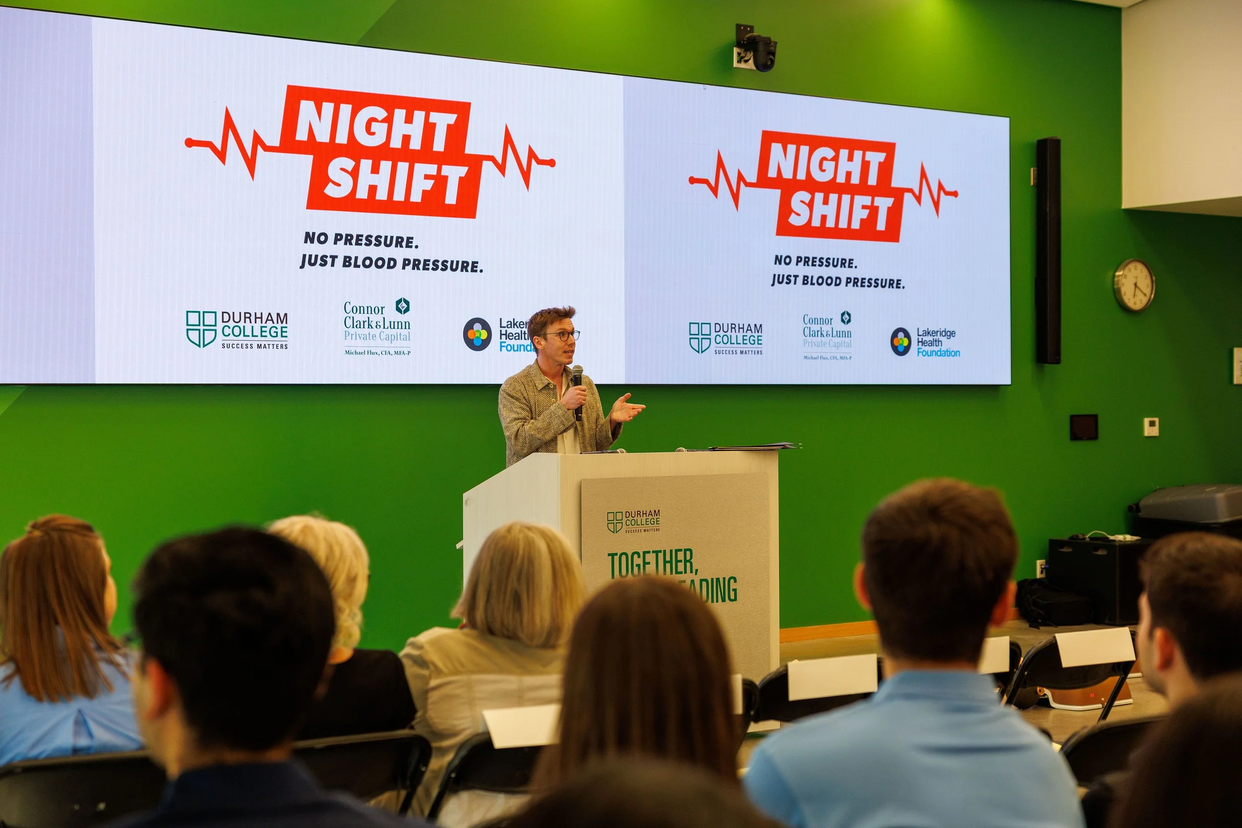 A man giving a presentation at Durham College titled 'Night Shift'. The slide on the screen has the words 'No Pressure. Just Blood Pressure.', with logos from Durham College, Connor Clark & Lunn Private Capital, and LakeRidge Health Foundation. Several audience members are seated, facing the speaker.