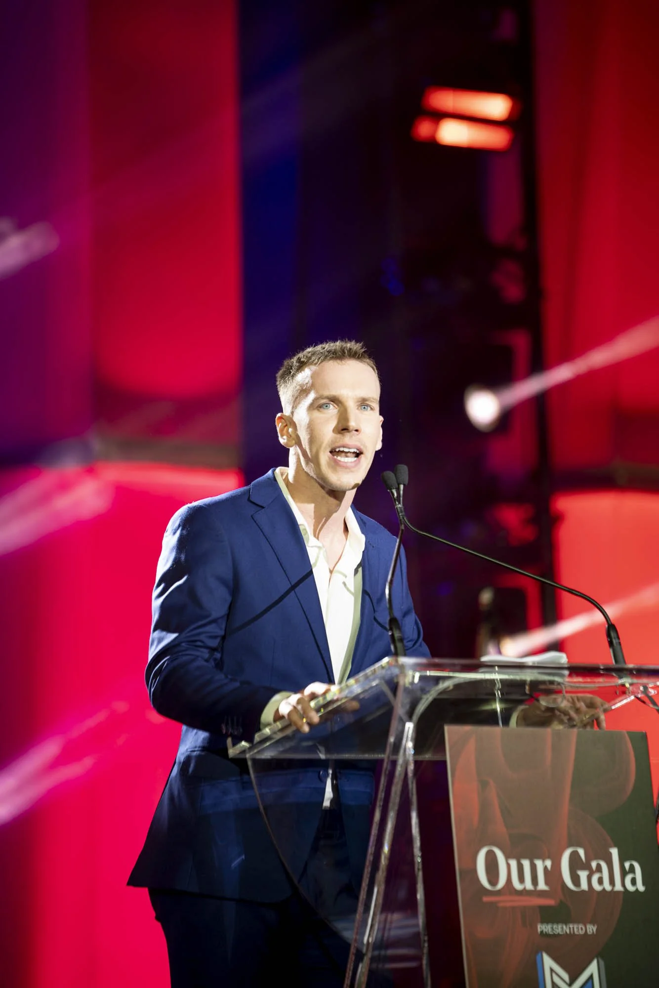 A man in a blue suit speaking at a podium with a sign that says 'Our Gala' during an event, with a red and purple background and stage lights.