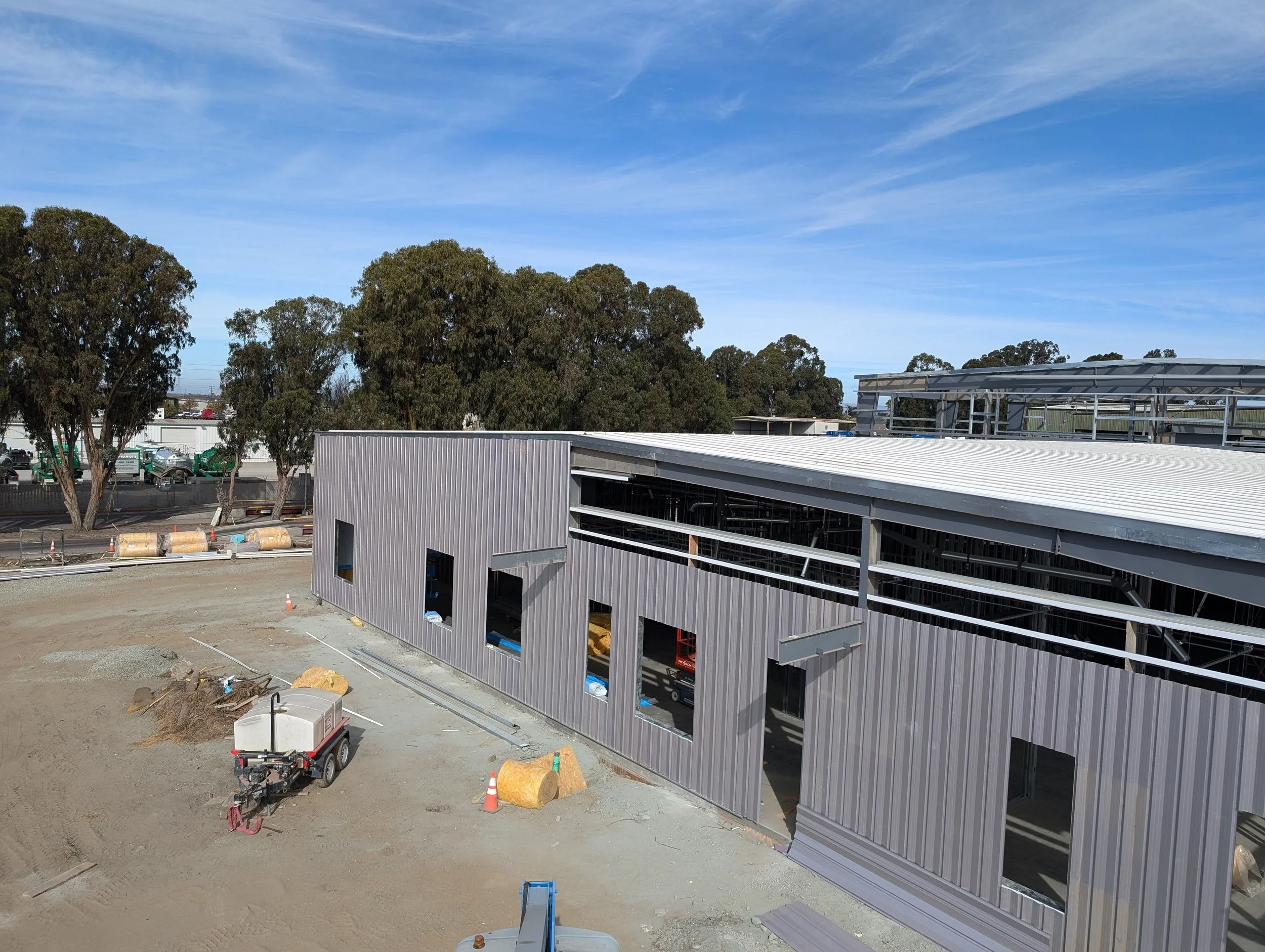 Construction site with a partially built metal building, construction materials, equipment, and large trees in the background under a blue sky with scattered clouds.