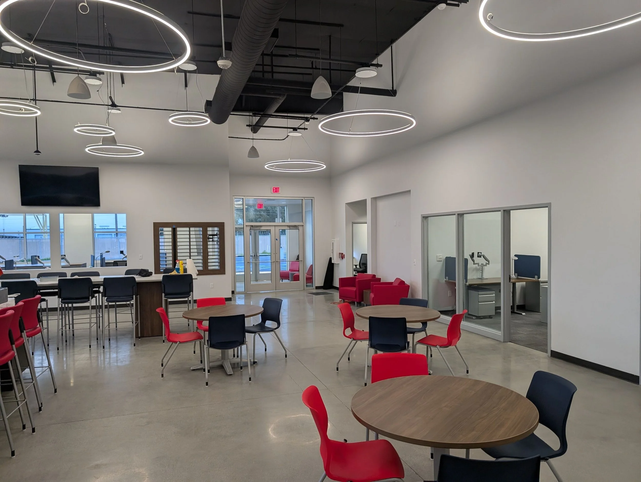 Modern lobby or break room with circular ceiling lights, tables with chairs in red and black, a reception counter, and small private rooms with equipment inside.