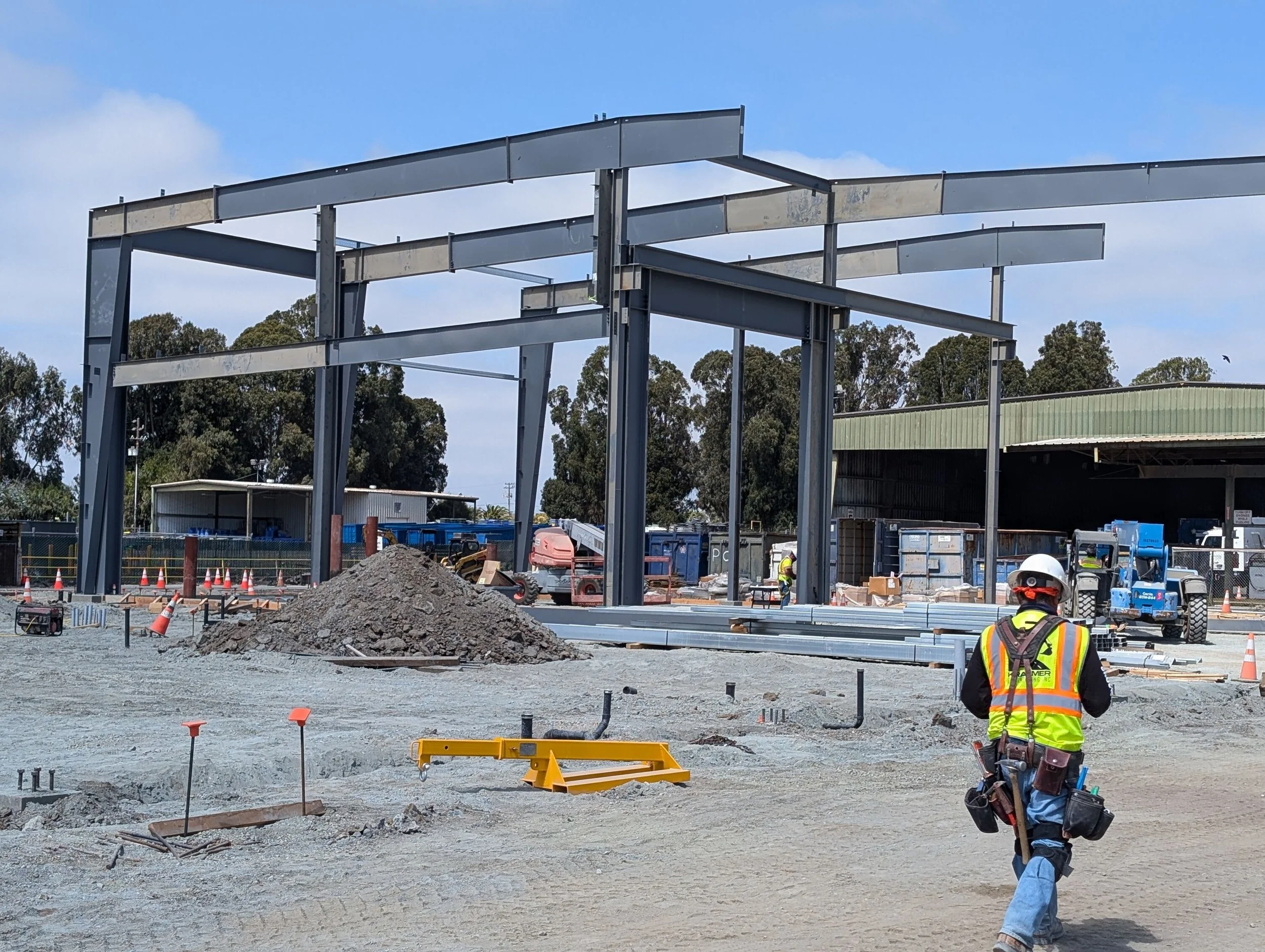 Construction site with steel framework being erected, piles of dirt, construction workers, and equipment.