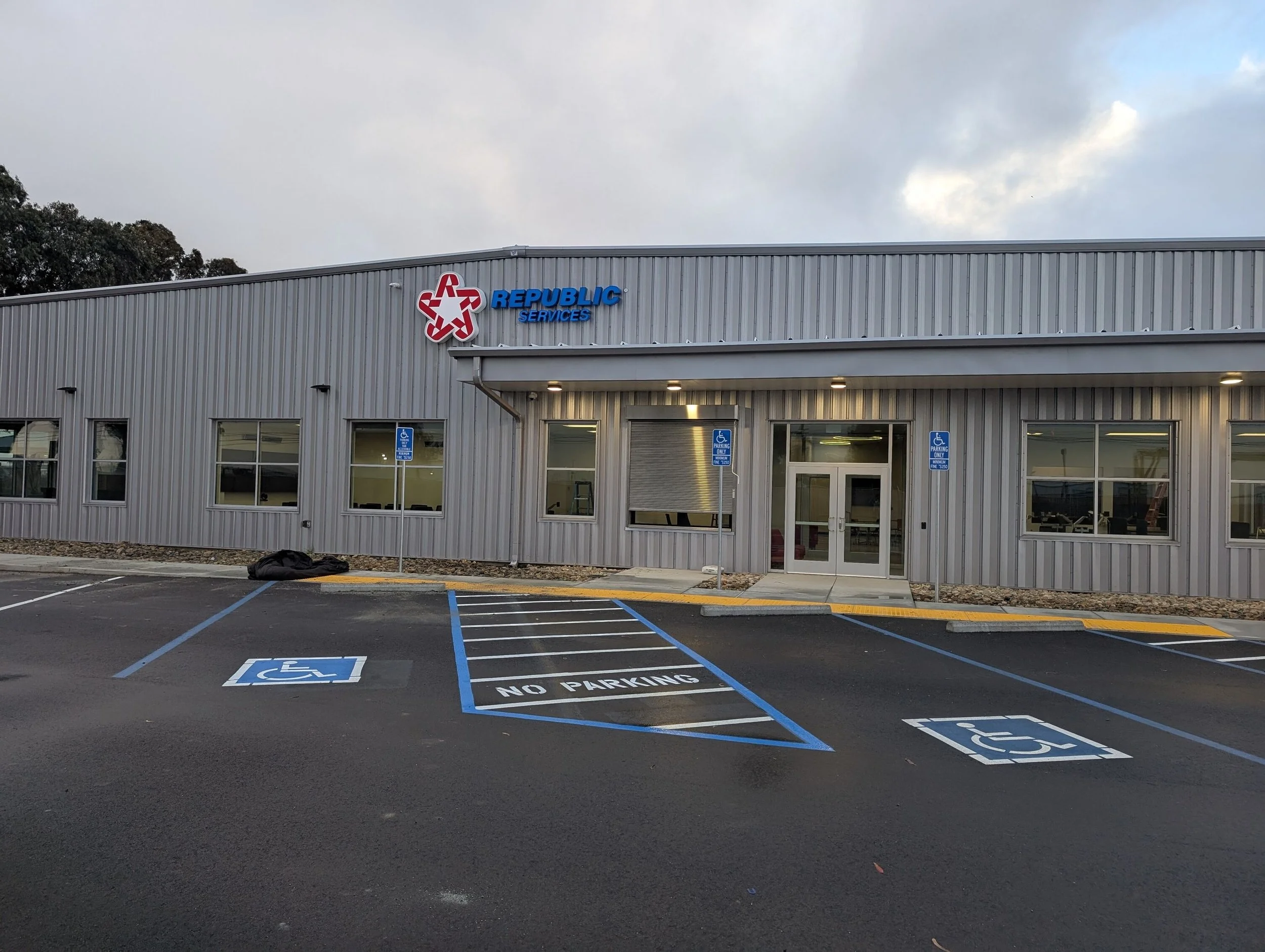 Exterior view of a commercial building with a sign reading 'REPUBLIC SERVICES'; the parking lot shows designated disability parking spaces with blue lines and signs, along with a no parking zone, and cloudy sky in the background.