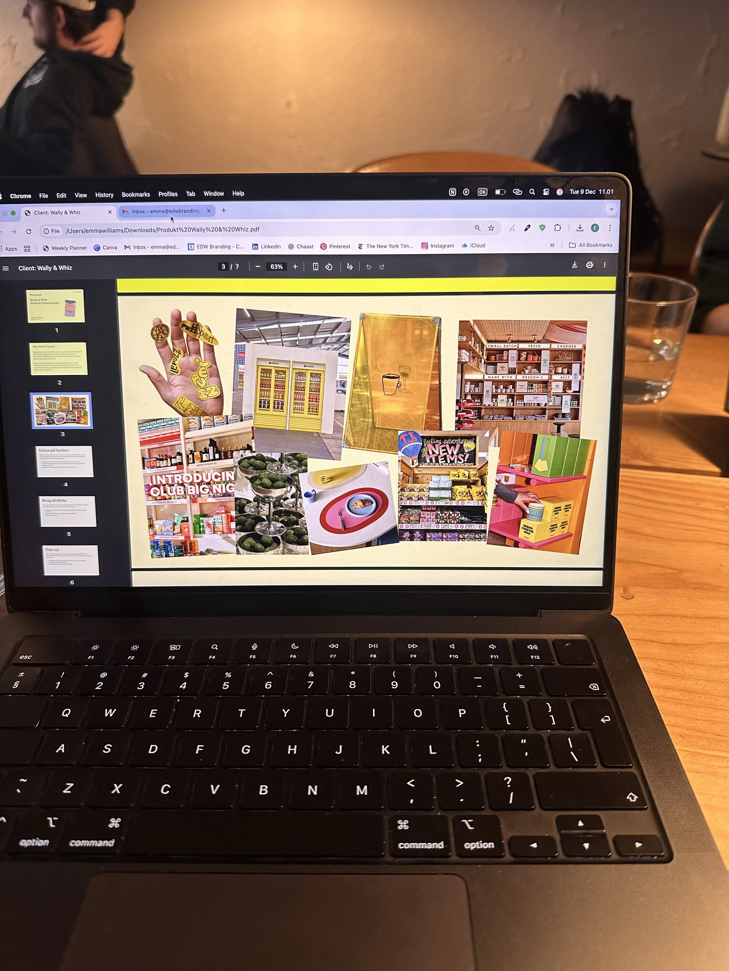 Laptop displaying a presentation with various colorful images of store shelves and products, on a wooden table in a cafe or similar setting, with a glass of water and a person in the background.