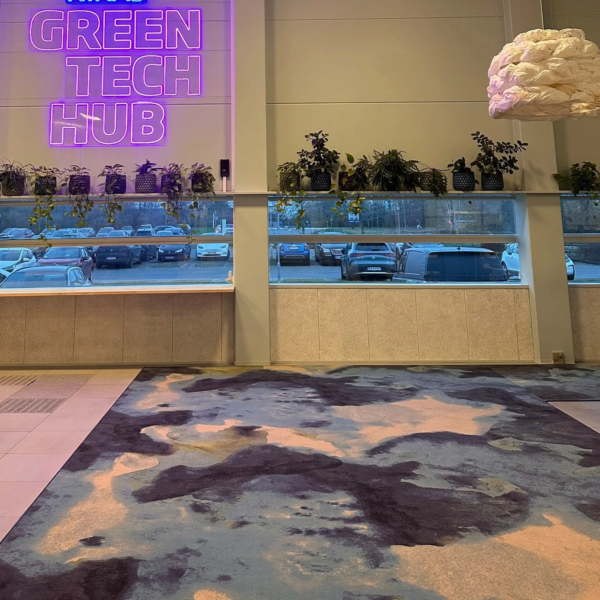 Interior view of the Green Tech Hub with a large neon sign, potted plants on a windowsill, and a colorful abstract carpet.