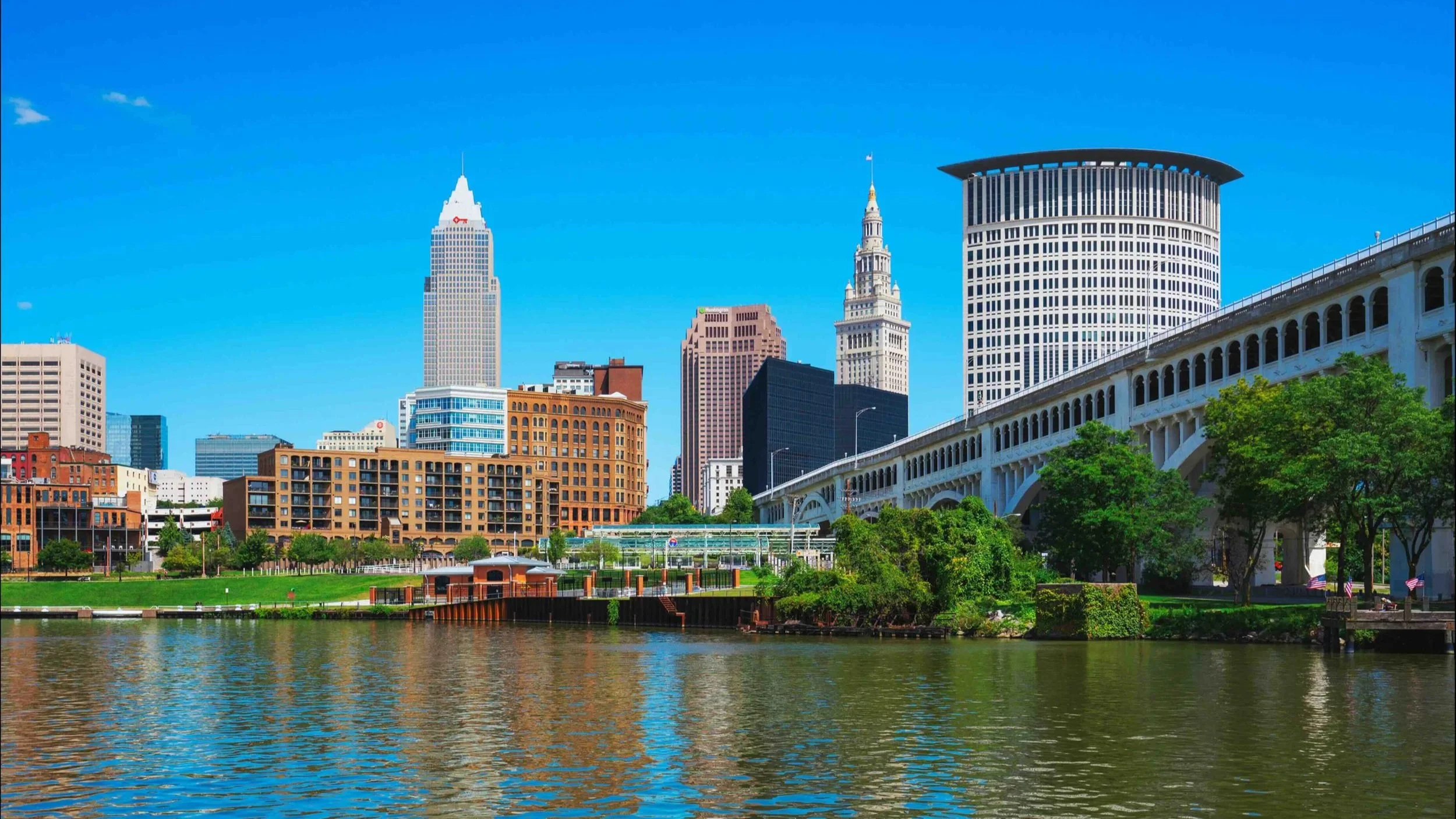 City skyline with tall buildings, a river in the foreground, greenery, and a clear blue sky.