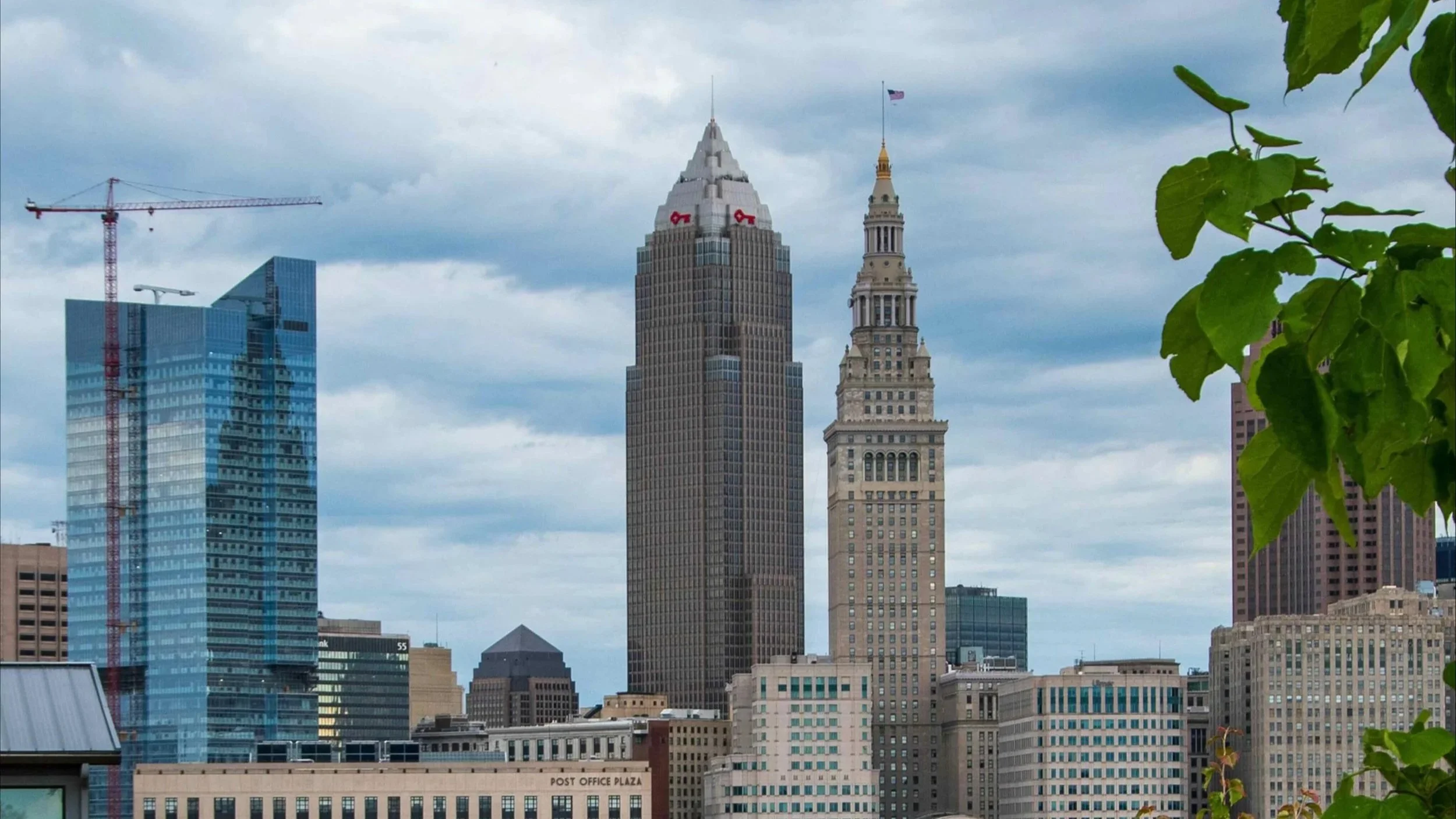City skyline with tall skyscrapers, including a modern blue glass building and the historic Flatiron Building, under a cloudy sky, with green leaves in the foreground.