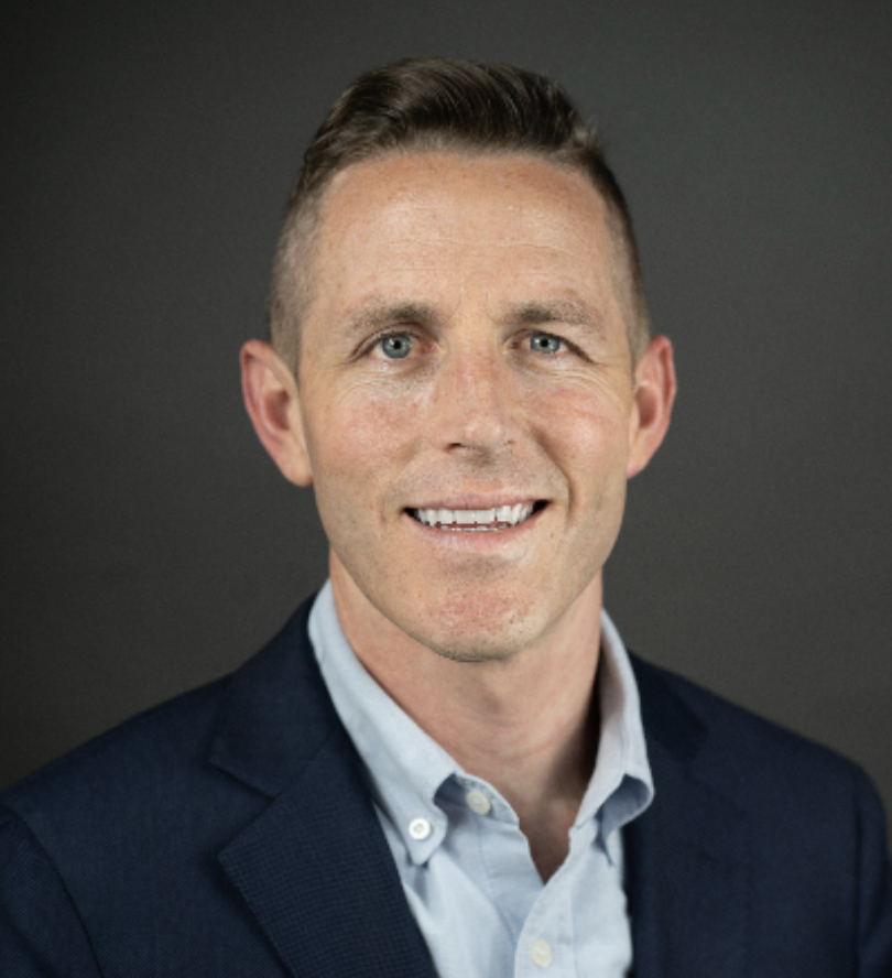 Professional headshot of a man with short brown hair, blue eyes, light skin, wearing a navy blazer over a light blue button-up shirt, smiling against a dark gray background.