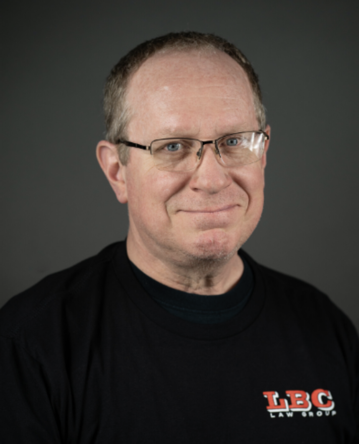 A middle-aged man with glasses, short light brown hair, wearing a black t-shirt with a red and white LBC Law Group logo, smiling against a dark gray background.