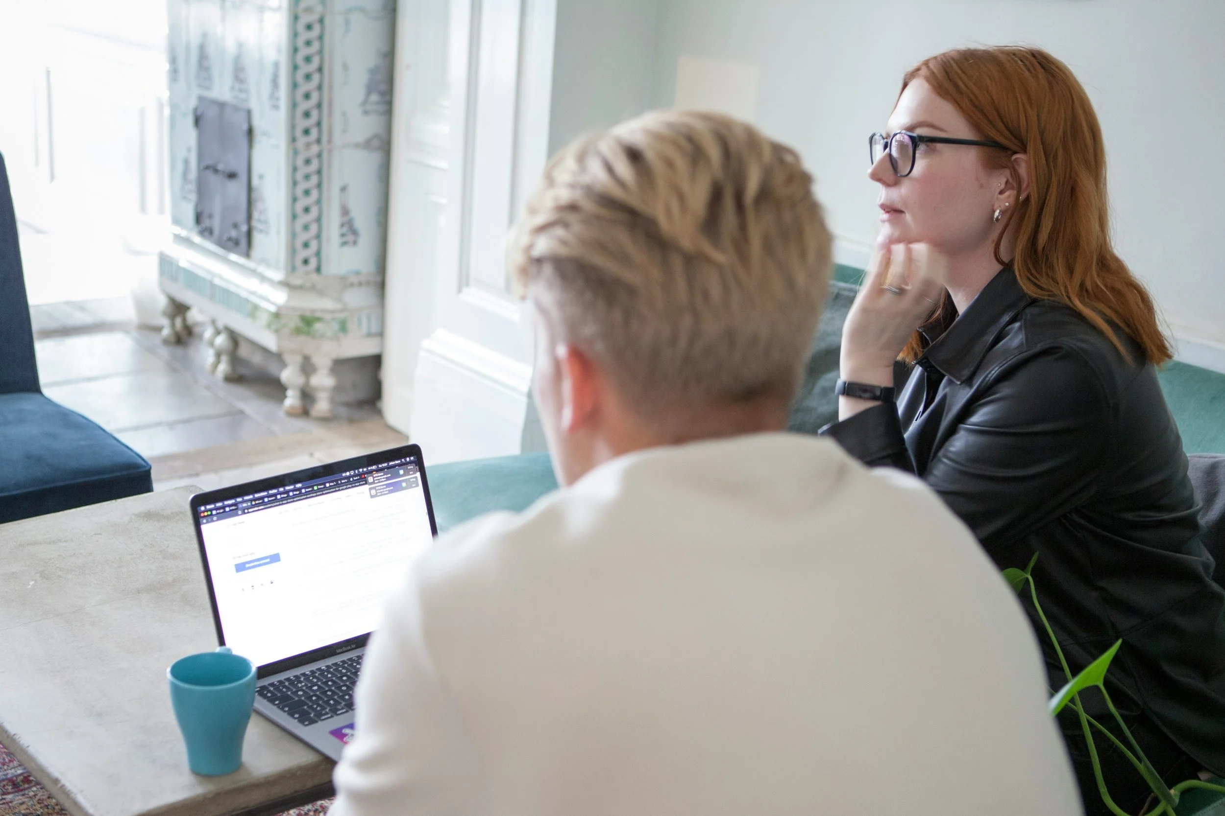 A man and a woman sitting at a table with a laptop and a blue cup, engaged in a discussion or meeting in a bright, modern room.